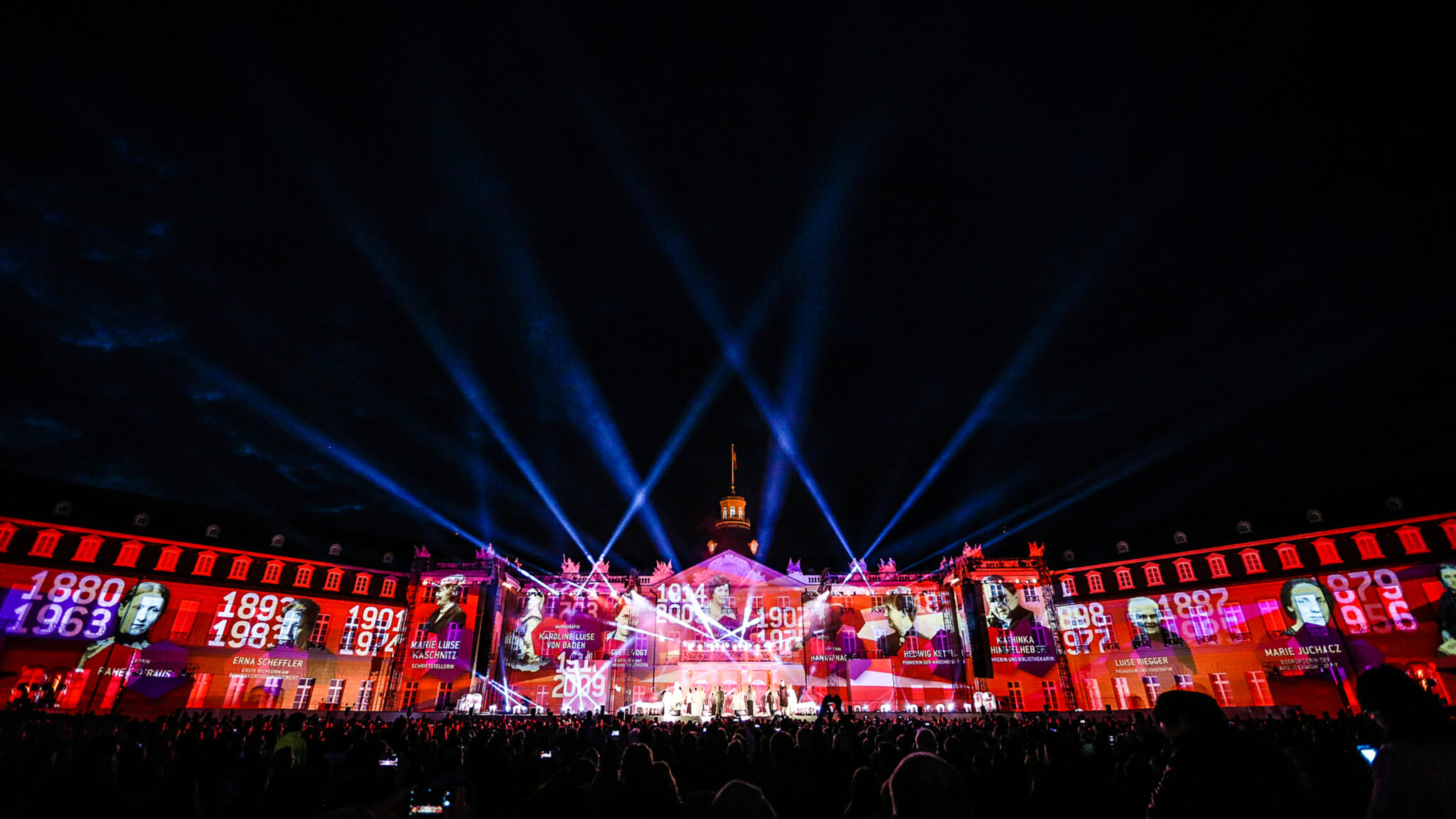 Blue searchlight beams fanning out over a palace façade covered in colourful projections, with an audience in the dark foreground.