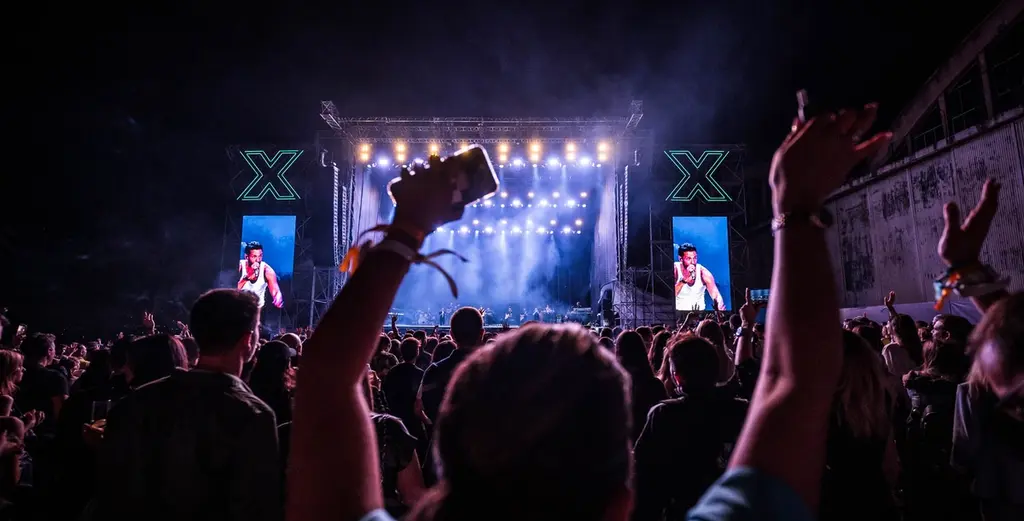 A large crowd enjoying a live concert at night, with vibrant stage lights and giant screens displaying the performer. Audience members raise their hands and phones in the air, capturing the energy of the moment. Battle Royal Studios