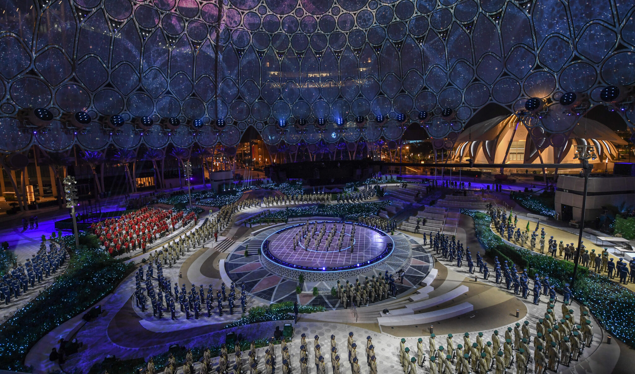 A large arena at night seen from above, blue projected light across the dome and floor and a dense crowd seated around a central stage.