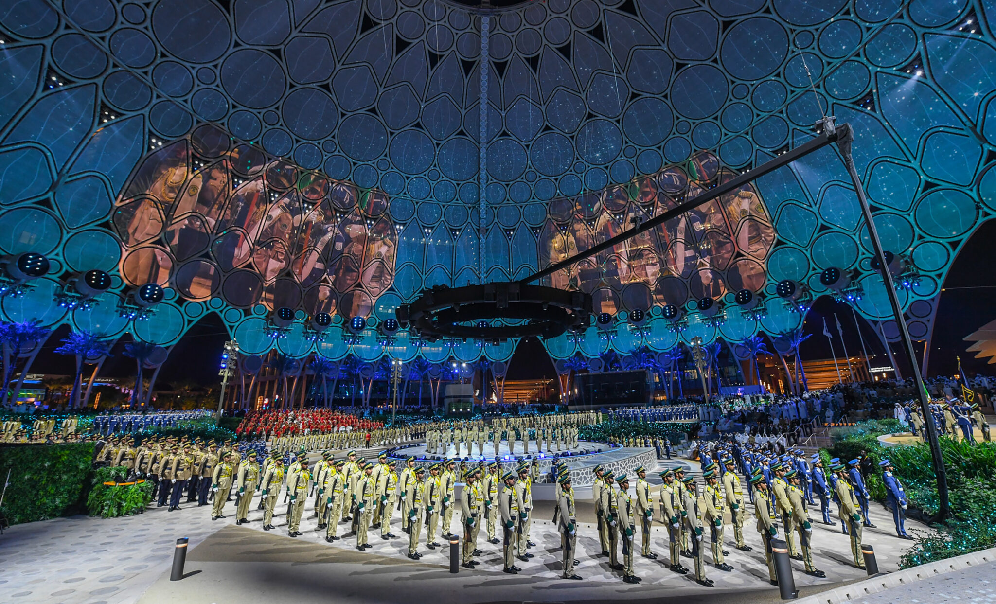 Wide view of a domed arena with a landscape projected across the ceiling and hundreds of performers arranged in formation on the floor.