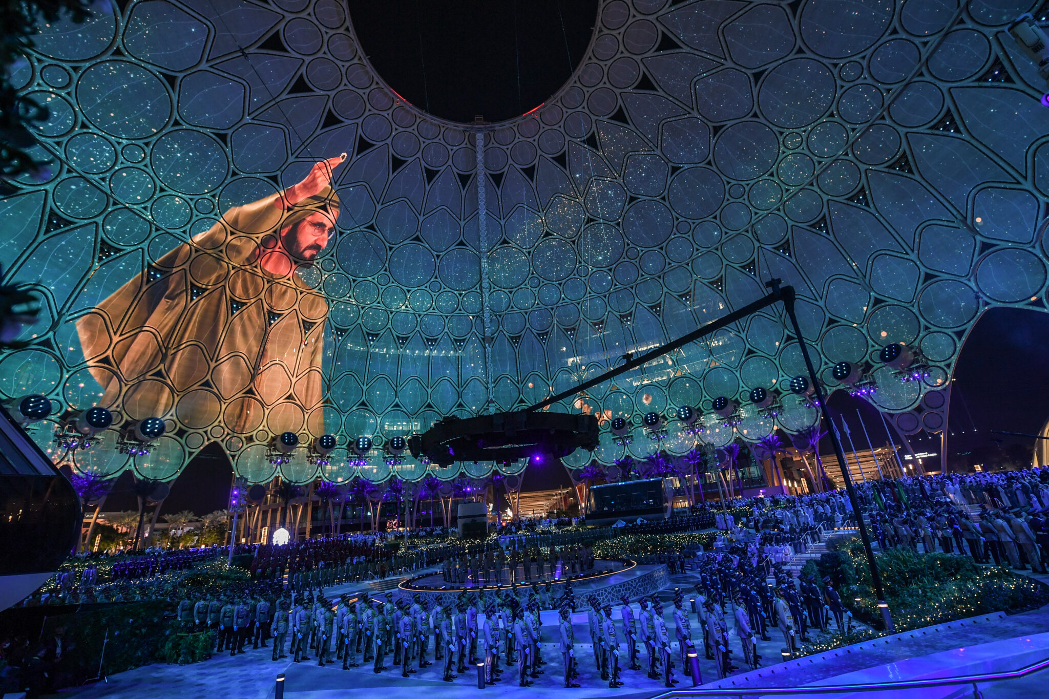 A tall robed figure in gold and cream stands against the curve of a teal-lit dome, with performers gathered on the floor below.