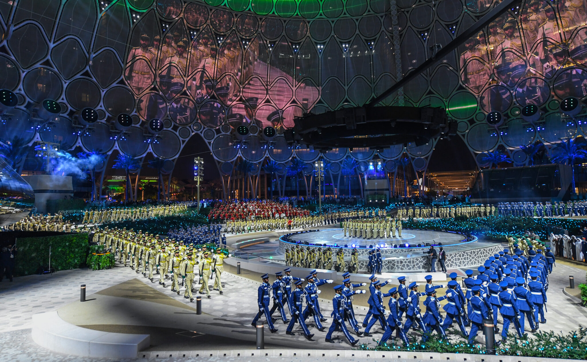 Performers in pale costume arranged in rows across a blue-lit arena floor, beneath a domed ceiling carrying projected imagery.