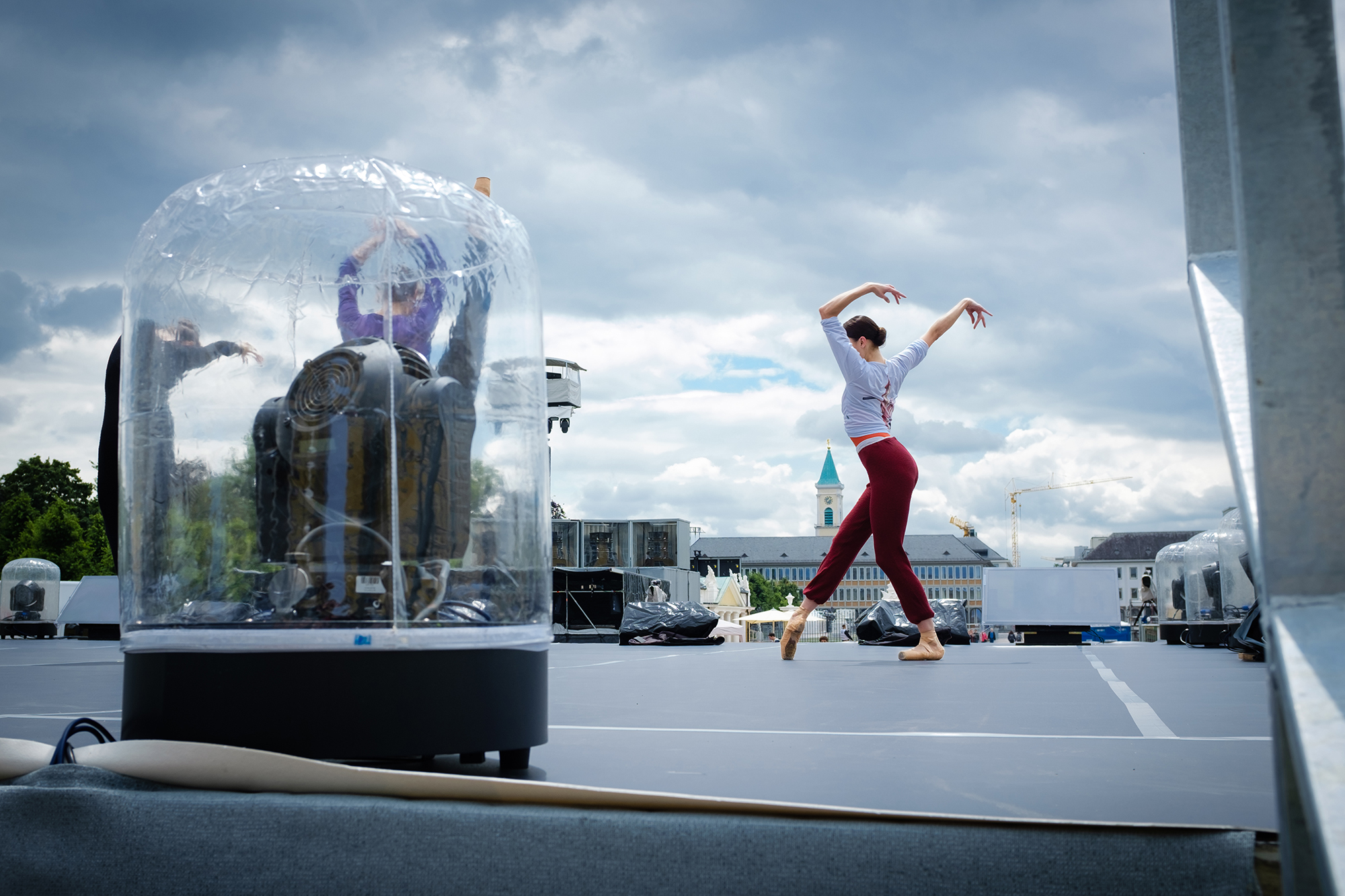 Daytime rehearsal on an outdoor stage: a dancer in red trousers poses with arms raised beside a large glass dome holding a seated costumed figure, under a cloudy sky.