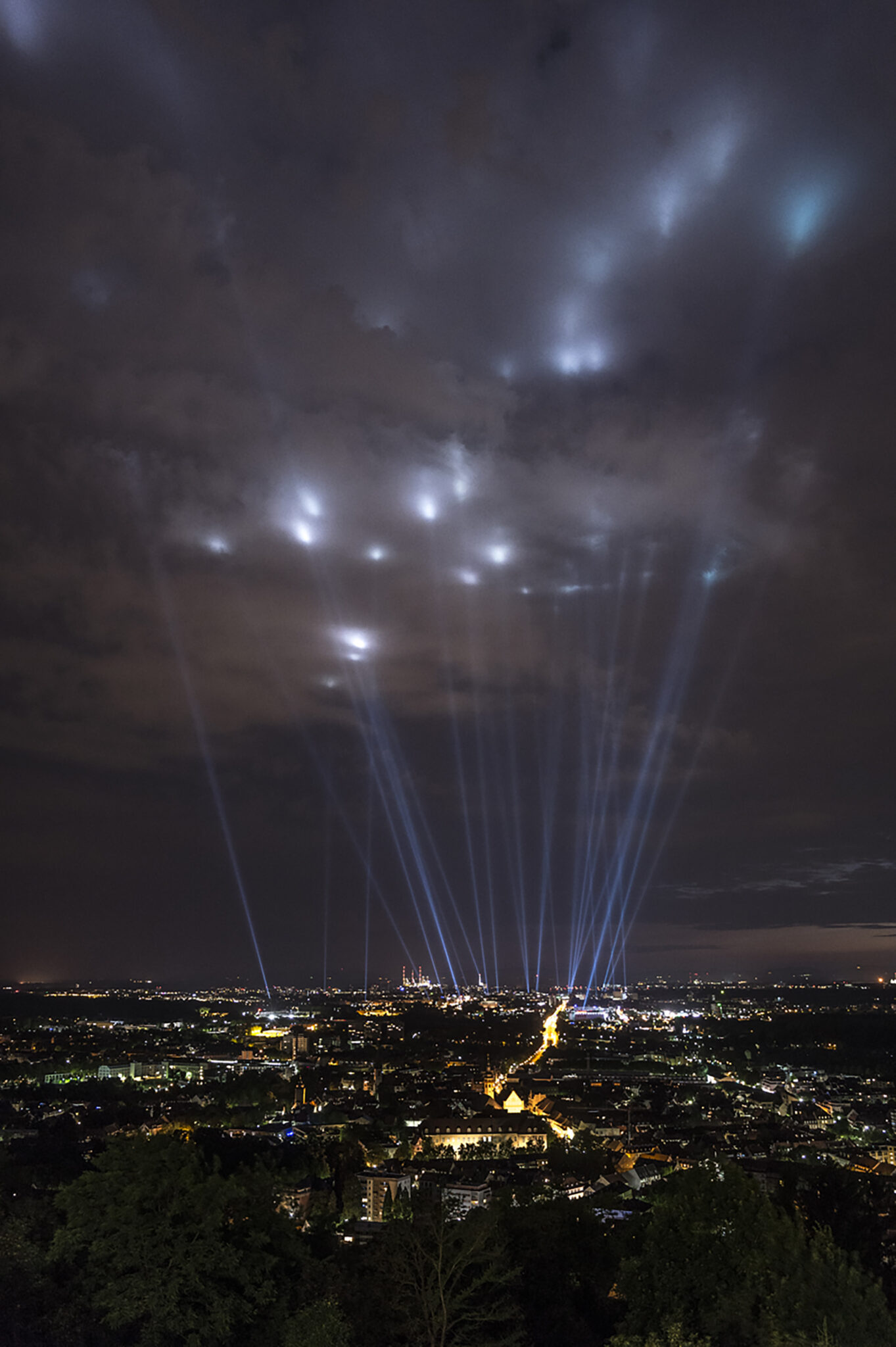 Elevated night view over city lights with bright searchlight beams shining upward into a cloudy sky.