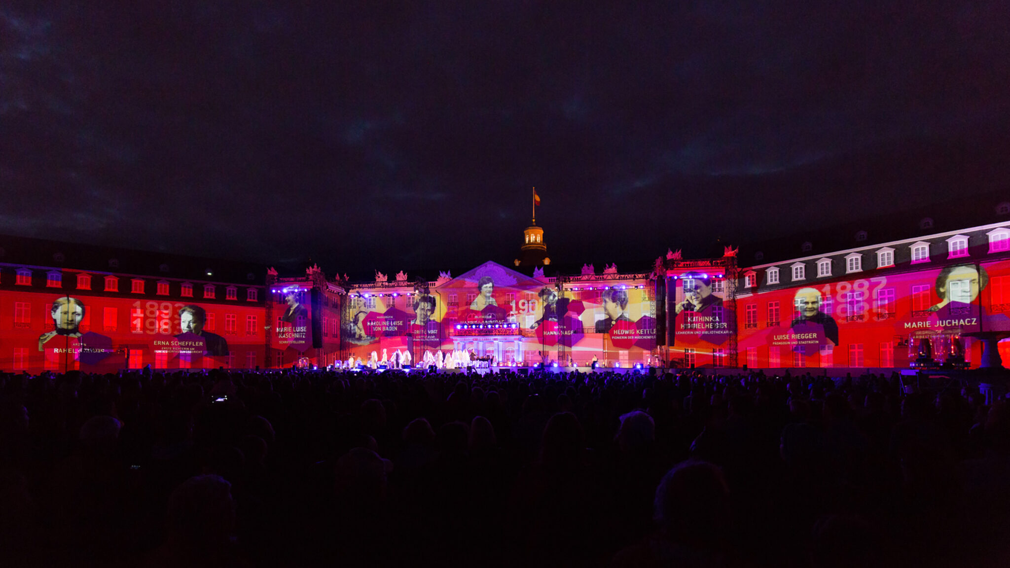Light projections and historical figures are displayed on a large building during a nighttime event with a crowd watching below. BattleRoyalStudios