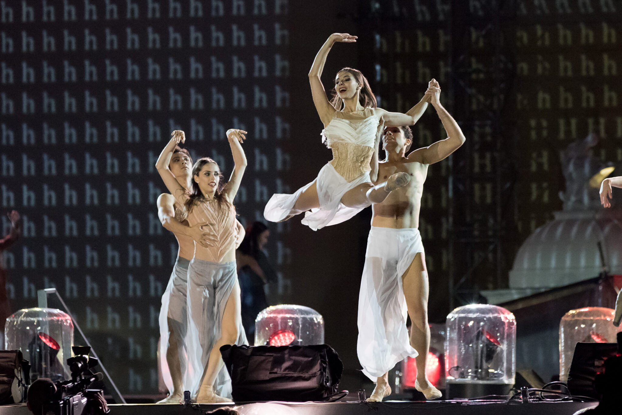 Three dancers in flowing white costumes performing on a dark stage, one lifted mid-leap, with glass domes and small lamps along the stage edge.