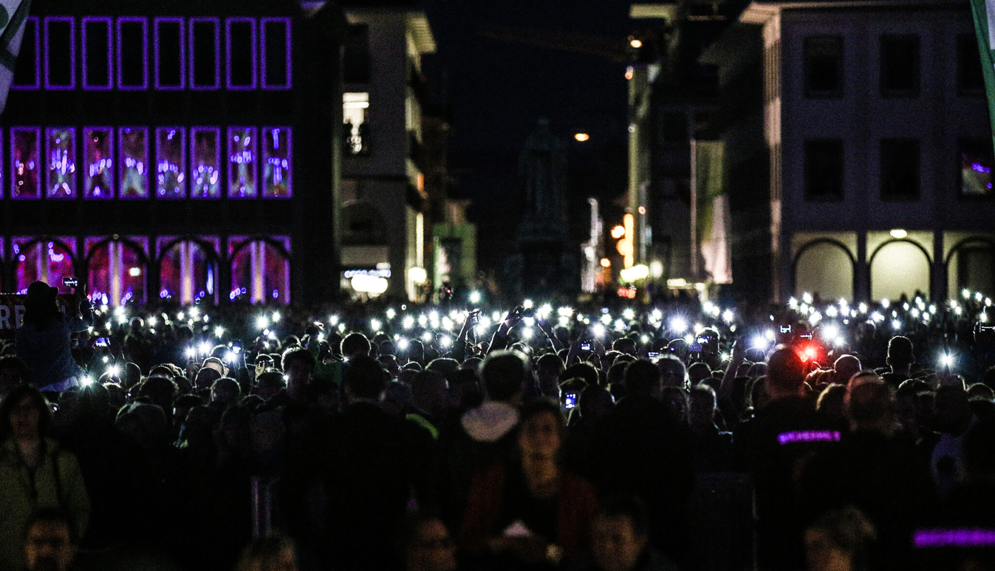 Nighttime crowd filling a city street, many holding up glowing phone lights, beneath building windows lit in purple.