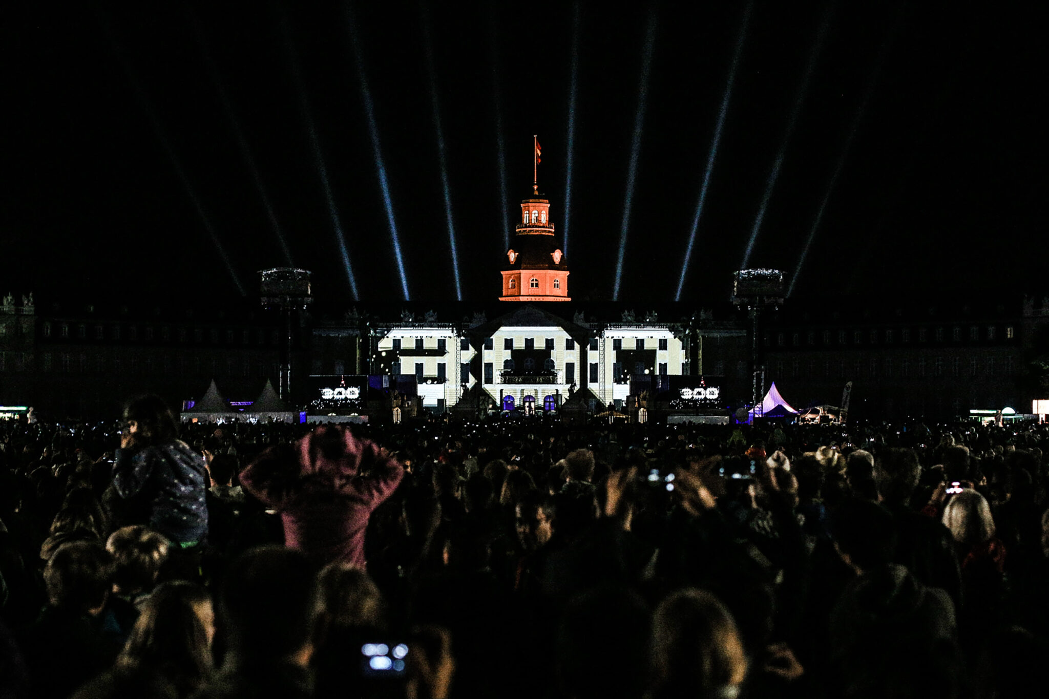 Large illuminated 300 letters in front of a palace tower at night, with searchlight beams rising into the sky above a dense crowd.