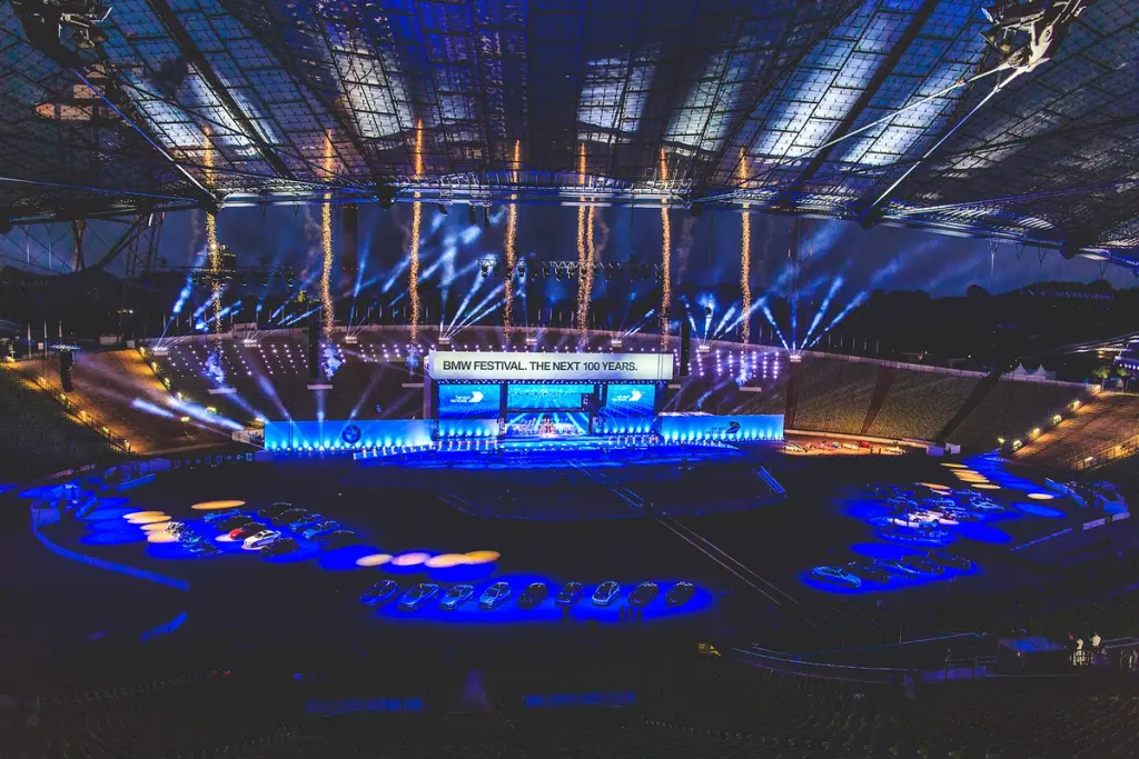 Panoramic view of the BMW Festival stage with dramatic lighting, fireworks, and cars on display in a stadium setting . Battle Royal Studios