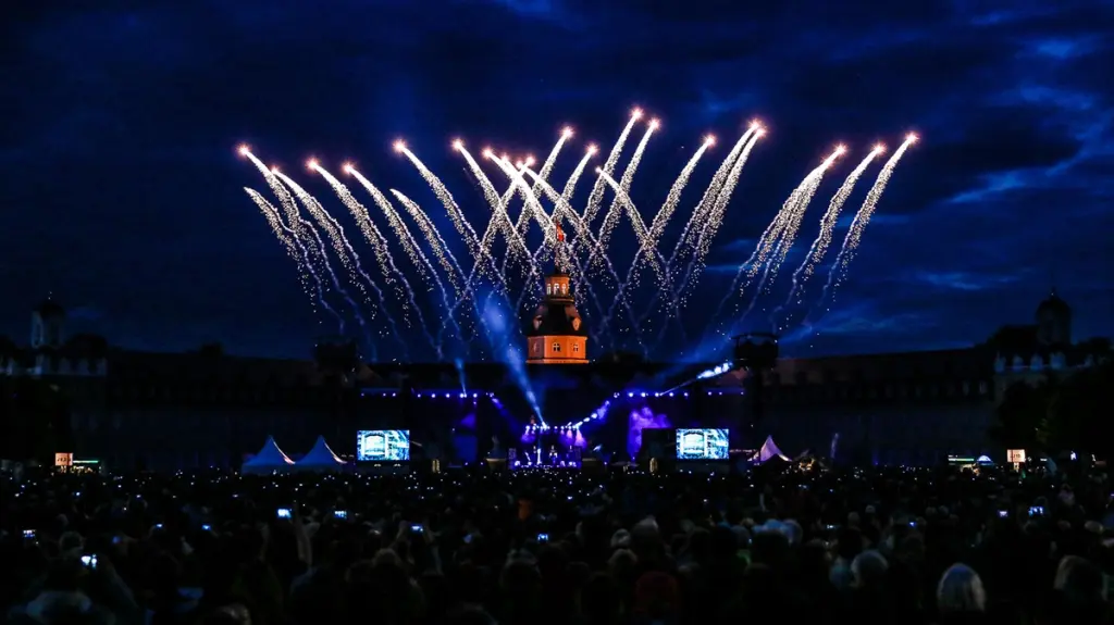 Fireworks light up the night sky over a large crowd and stage during an outdoor event. BattleRoyalStudios