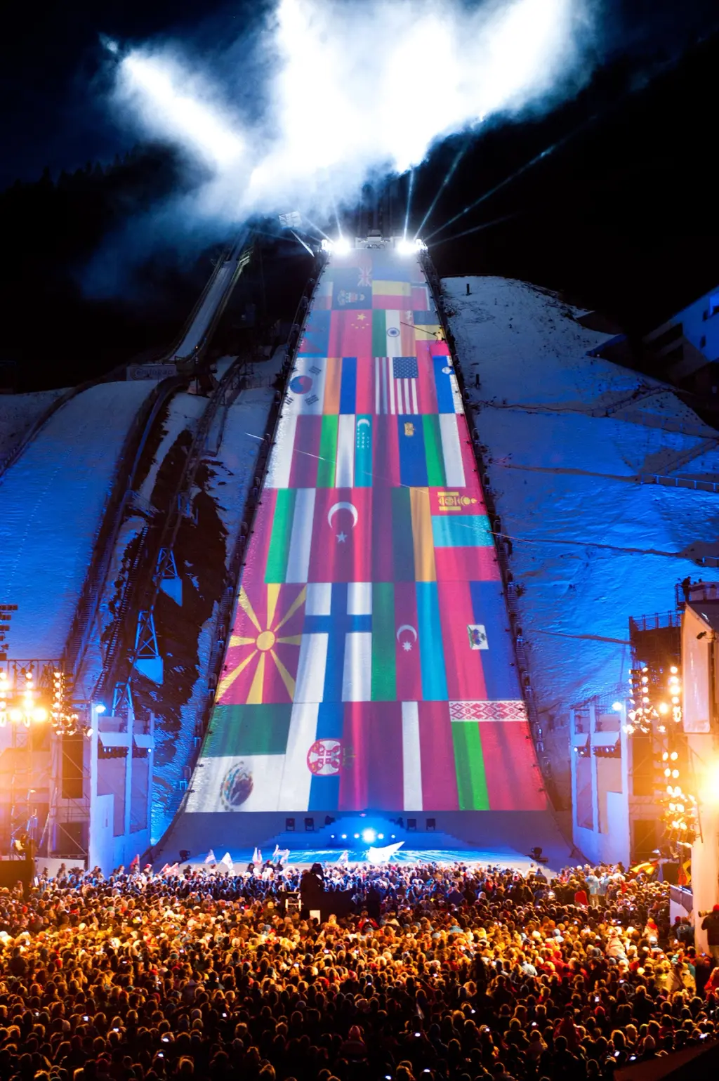 Large outdoor event with international flags projected onto a ski jump structure, illuminated as a crowd gathers below. BattleRoyalStudios