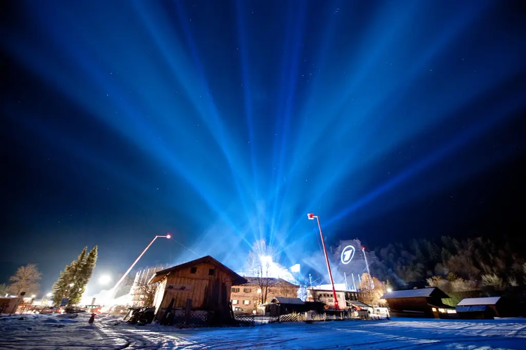 Bright blue lights fan out into the night sky over a snow-covered rural scene with wooden houses and event structures. Equipment and lights are visible, suggesting a large outdoor setup or event. BattleRoyalStudios