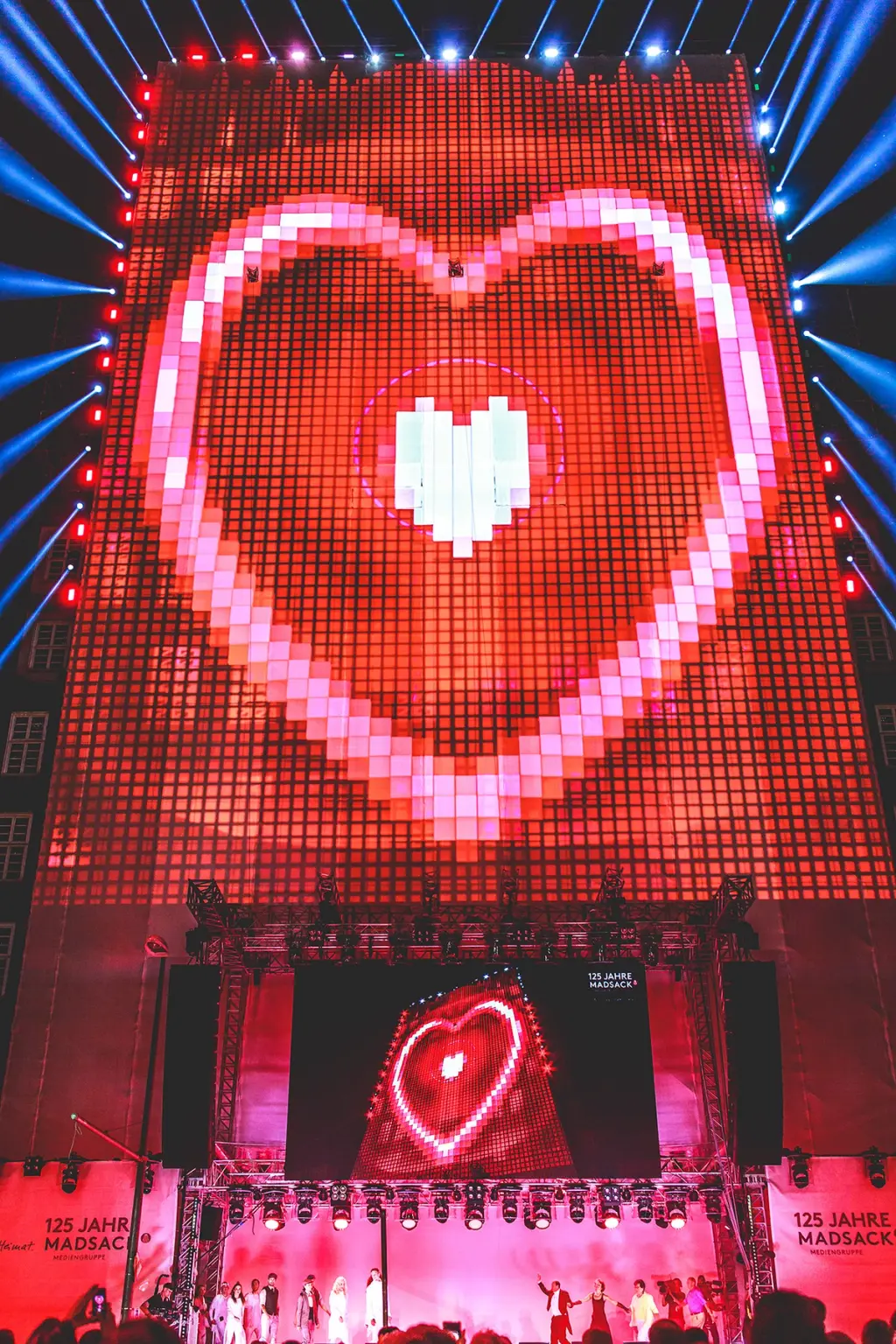 Massive LED screen displaying a large pixelated red heart with white and pink accents during a live event. Performers stand on stage below, celebrating "125 Jahre Madsack Mediengruppe." BattleRoyalStudios