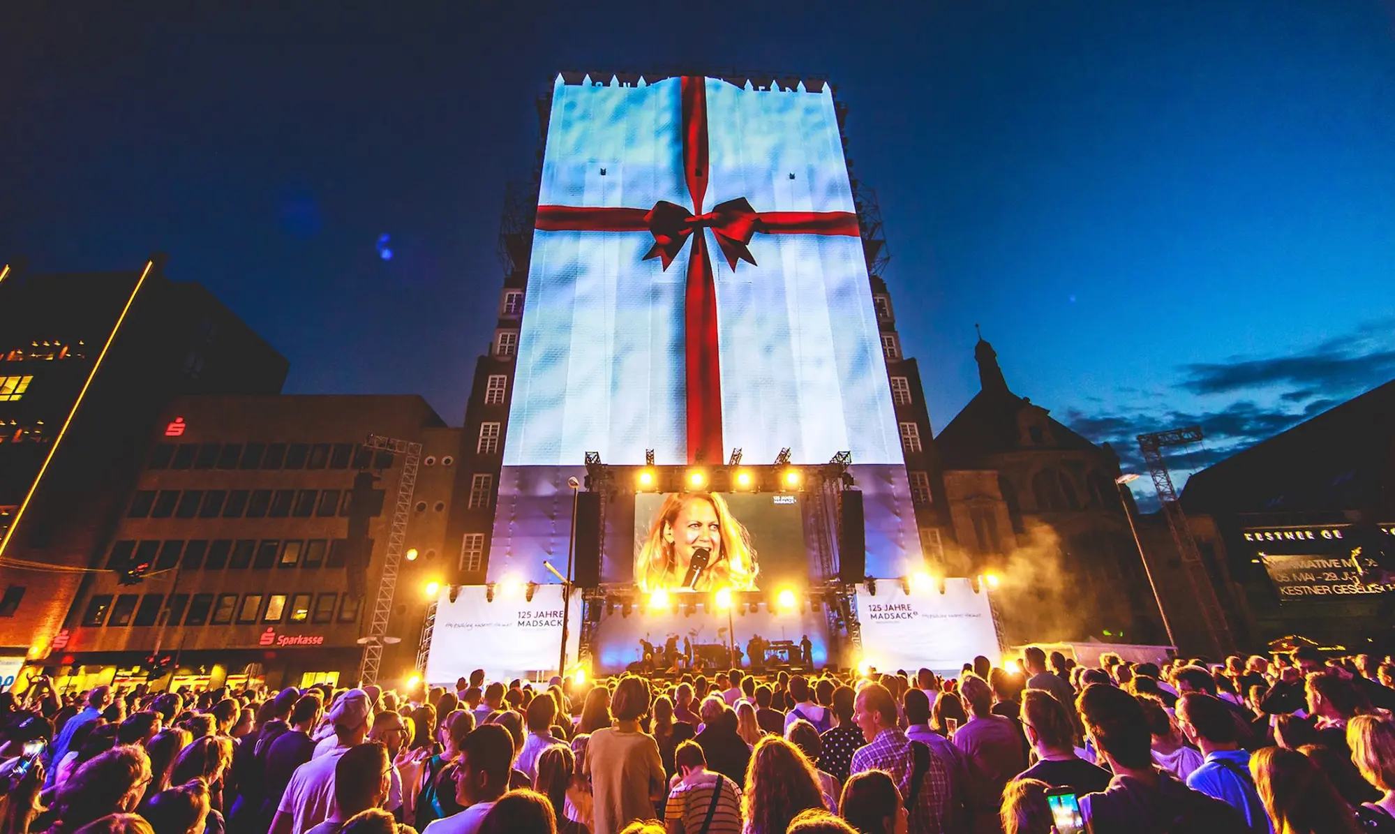 A large crowd gathers for an outdoor event in front of a building wrapped in a giant projection of a gift box with a red ribbon, while a singer performs on stage below. Battle Royal Studios