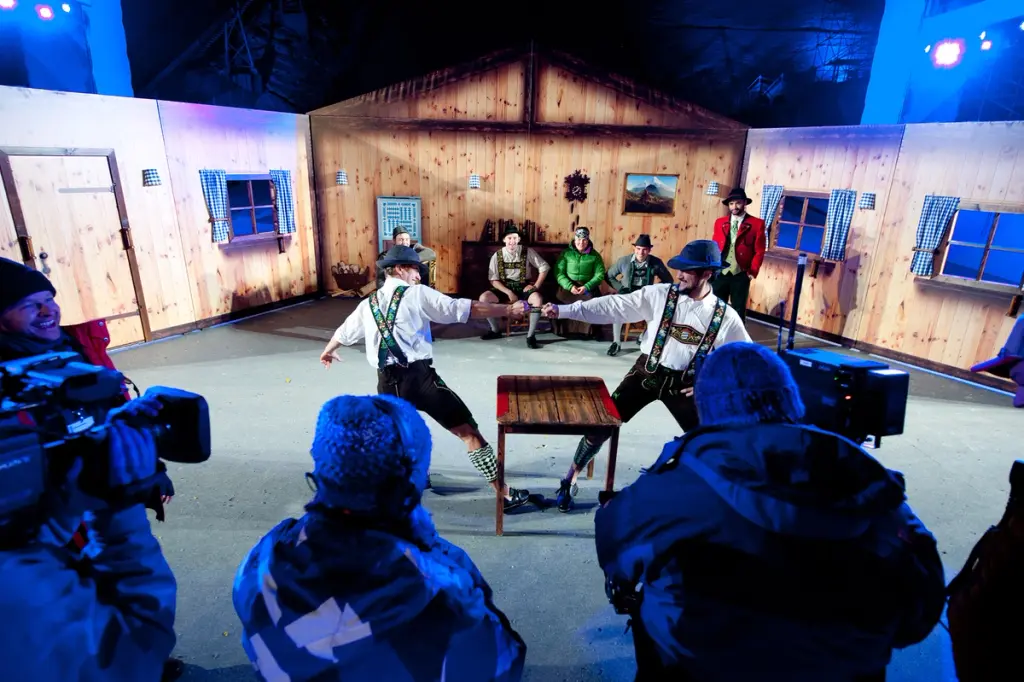 Two men in traditional Bavarian clothing engage in a finger-wrestling contest, being filmed in a set resembling a wooden alpine cabin. Spectators and a film crew observe the scene. BattleRoyalStudios