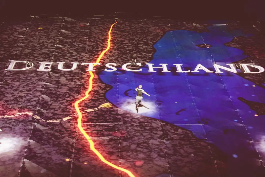 A performer walking on a large map projection with the word "Deutschland" prominently displayed, highlighting Germany with a red line marking the border. Battle Royal Studios