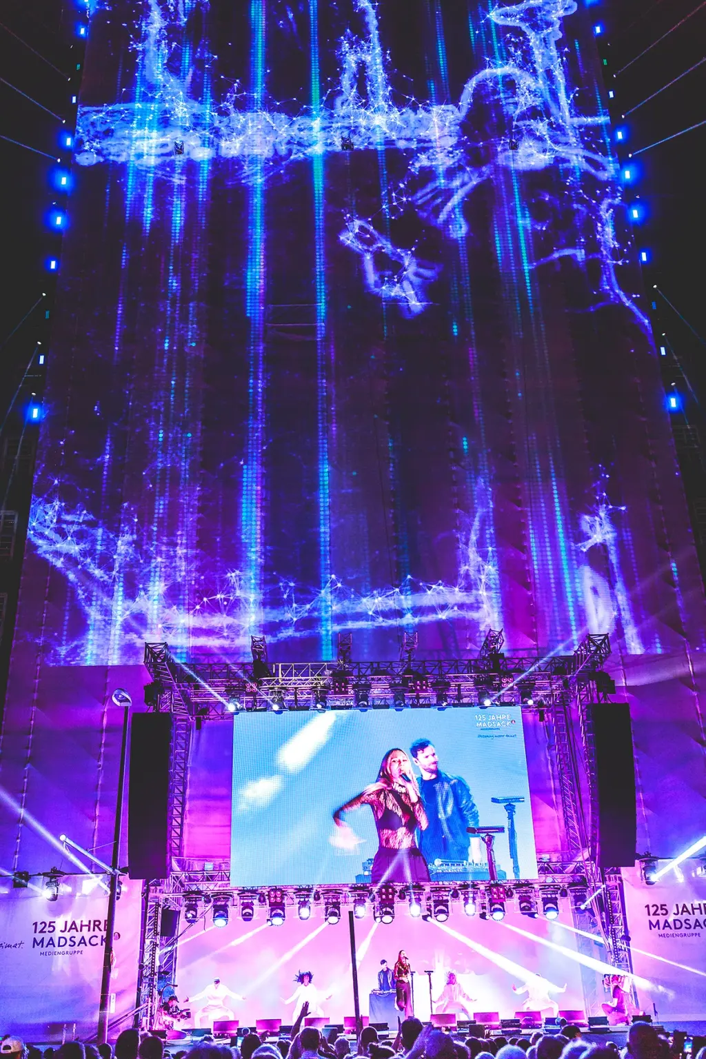 A large outdoor stage with a performer singing in front of a massive, illuminated screen. The screen displays vibrant blue and purple visuals that extend high into the sky, creating a dramatic backdrop for the performance. The event is part of the "125 Jahre Madsack" celebration. Battle Royal Studios