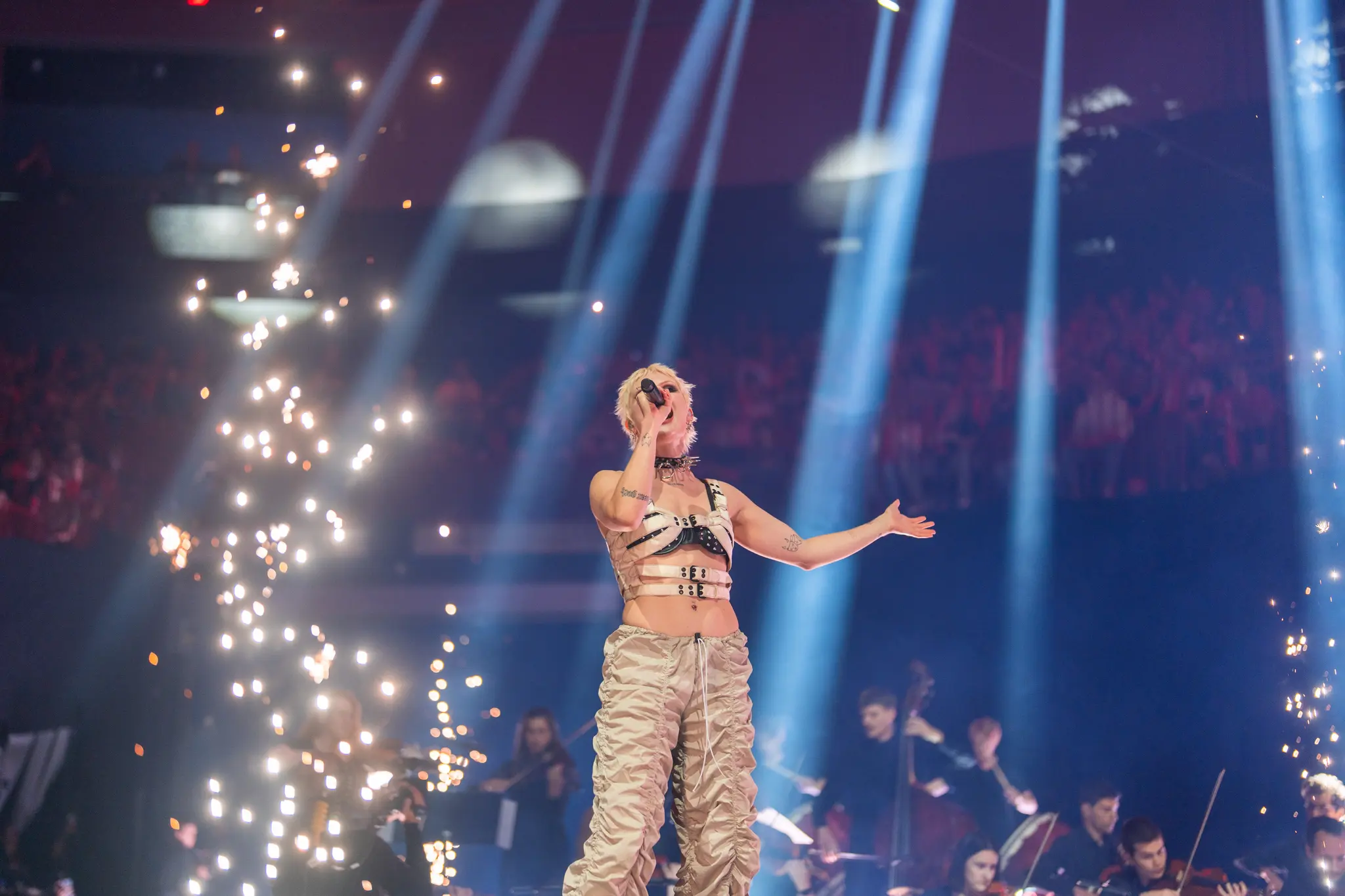 A singer performs on stage, holding a microphone and surrounded by dramatic spotlights and sparks, with an orchestra playing in the background. Battle Royal Studios