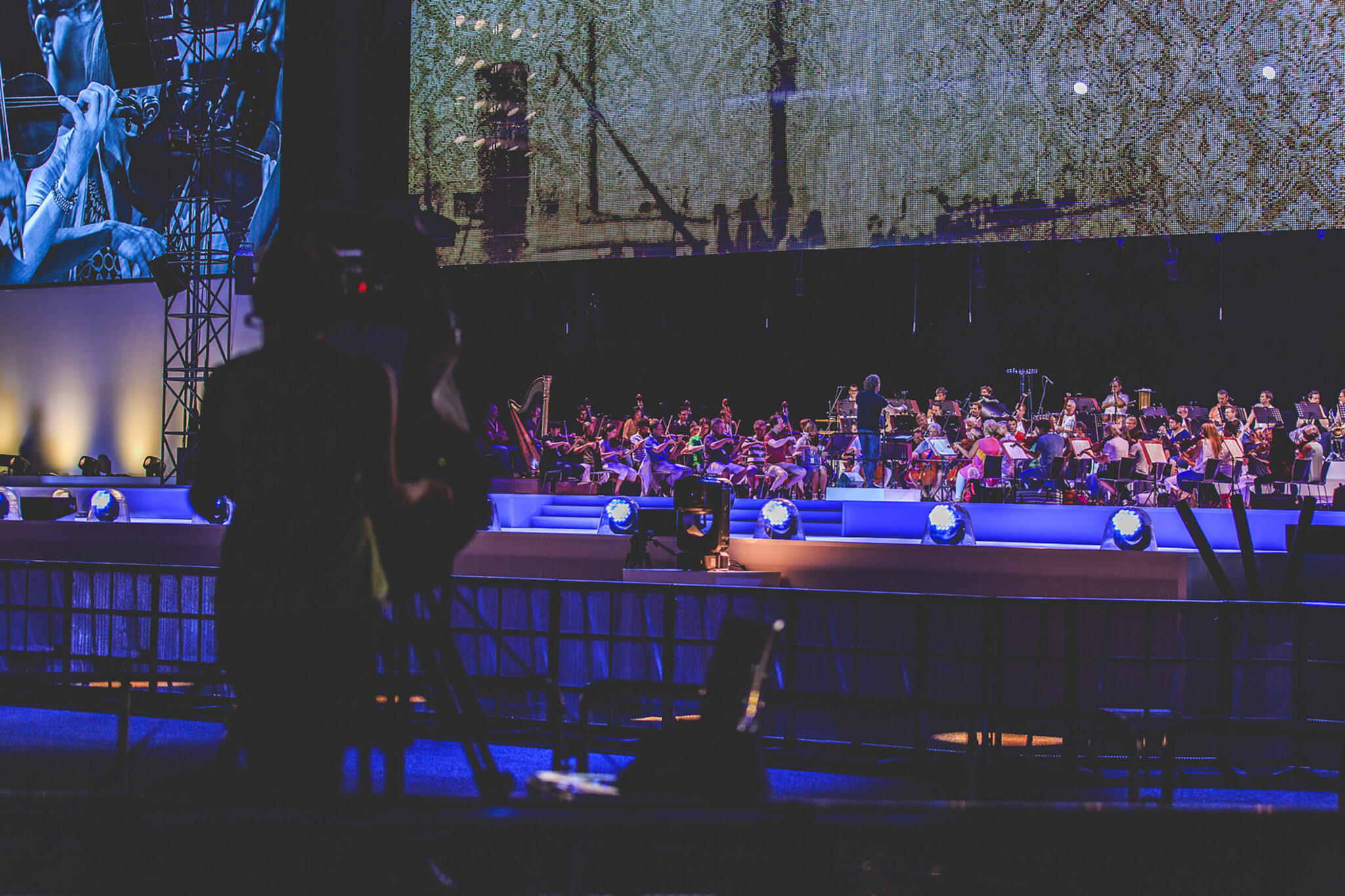 Orchestra performing on stage with a large screen displaying close-up images of musicians at the BMW Festival. Battle Royal Studios