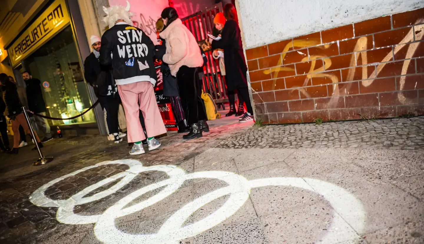 People stand outside an urban venue, with one person wearing a jacket that says "Stay Weird," while an illuminated Audi logo is projected onto the sidewalk. Battle Royal Studios