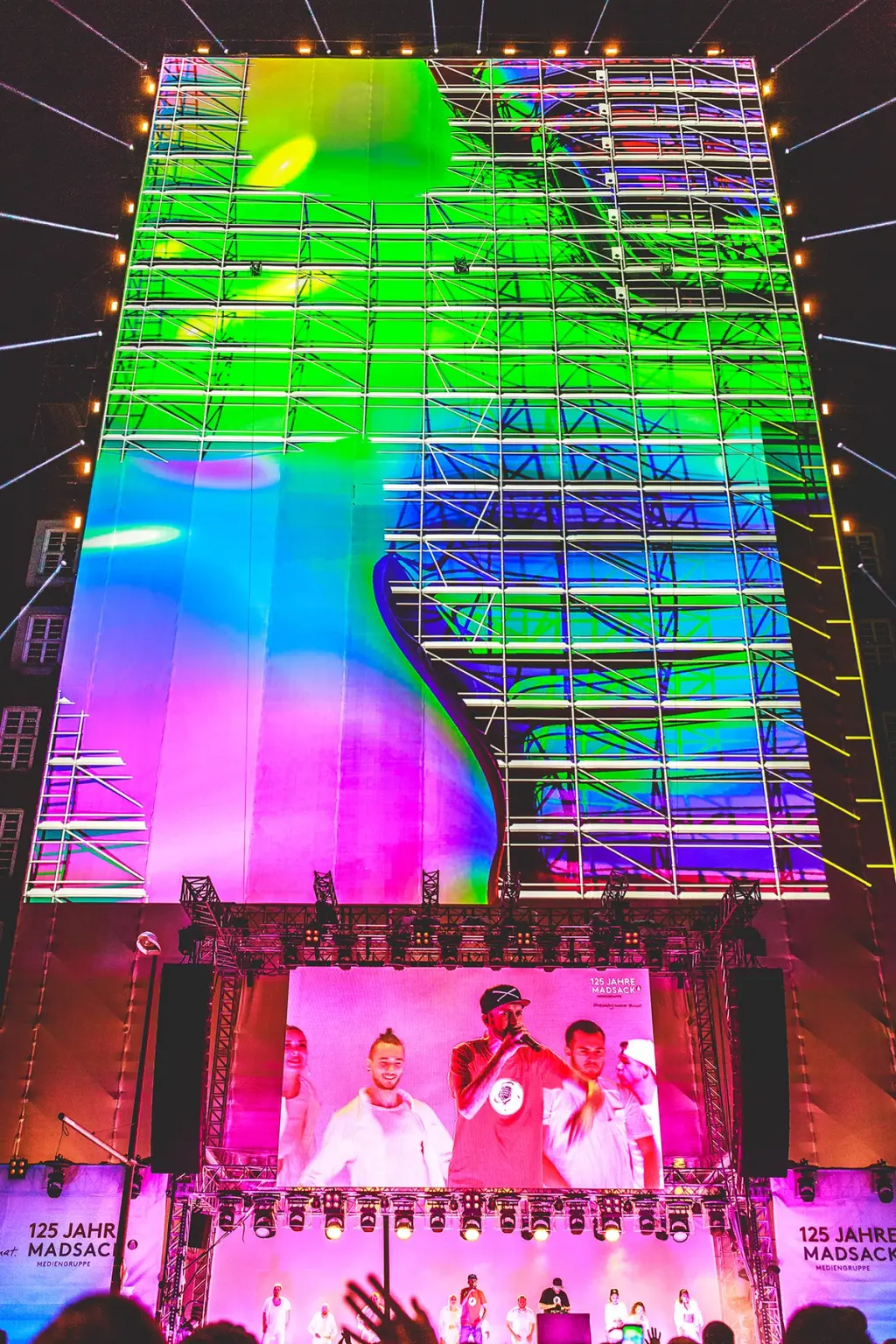 Giant LED screen displaying vibrant colors and abstract visuals during a live performance with a rapper in a red shirt on stage. The event is celebrating "125 Jahre Madsack Mediengruppe."  Battle Royal Studios