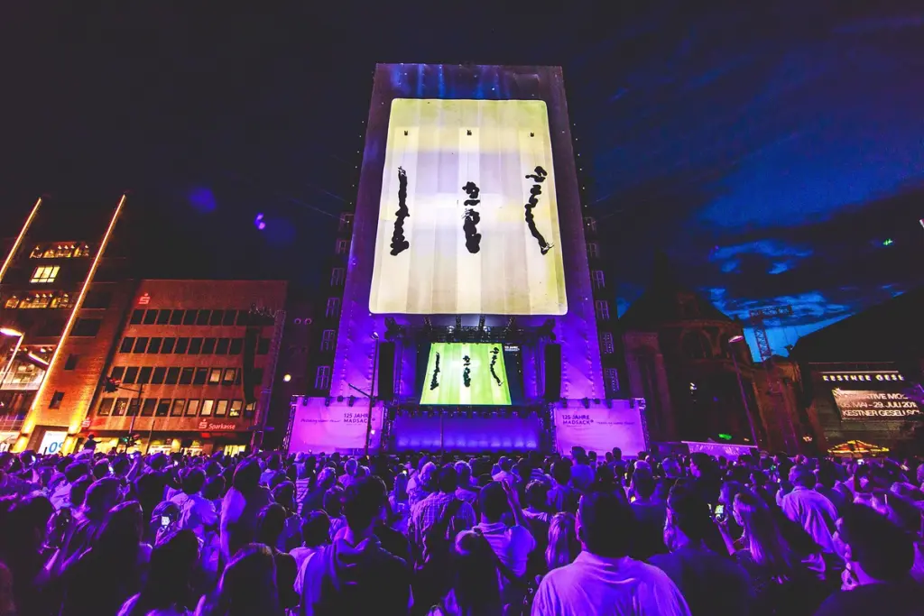 Large crowd gathered in a city square at night, watching a dynamic projection on a tall building during a public event or festival. Battle Royal Studios