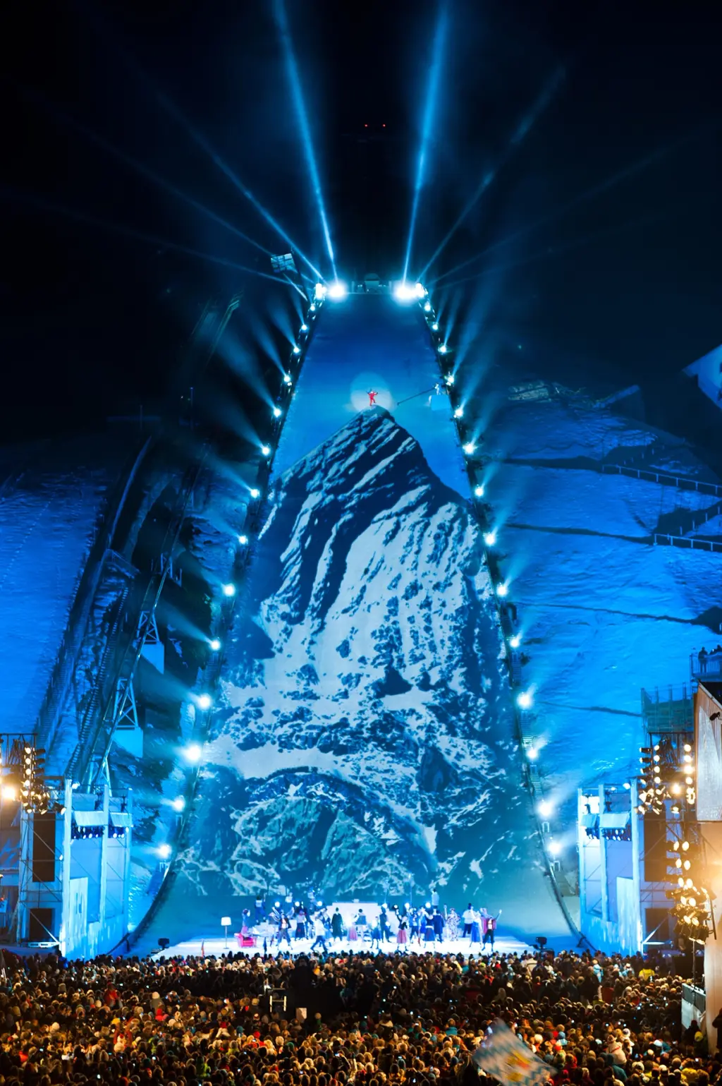 Large outdoor event with a projected snowy mountain on a ski jump structure, illuminated in blue, as a crowd watches below. BattleRoyalStudios