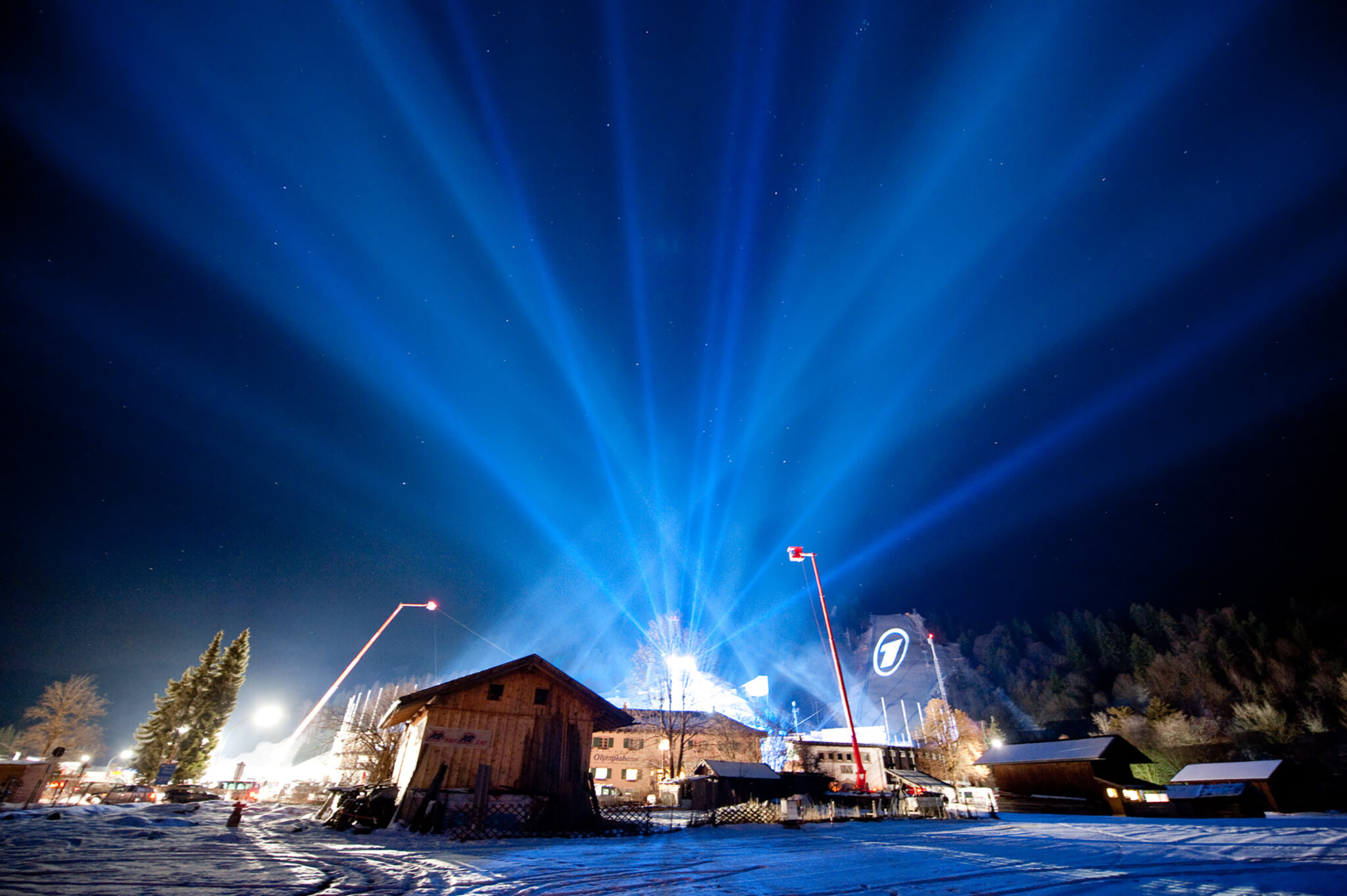 Bright blue lights fan out into the night sky over a snow-covered rural scene with wooden houses and event structures. Equipment and lights are visible, suggesting a large outdoor setup or event. BattleRoyalStudios