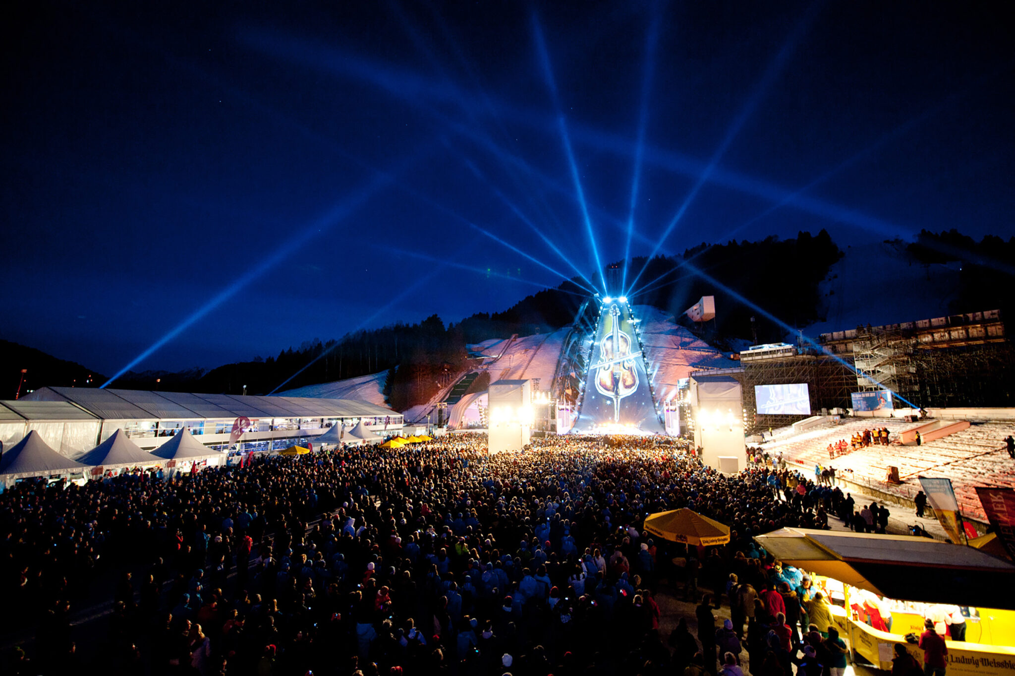 Wide night view of a large crowd in a snowy stadium, with blue light beams radiating from an illuminated stage at the base of a ski jump, and tents and screens along the sides.