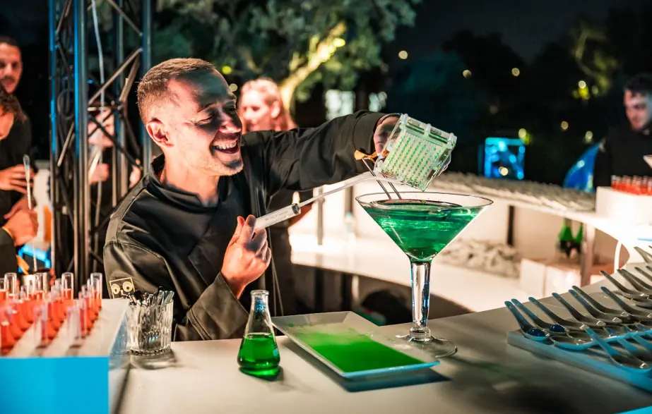 A smiling bartender pours a bright green liquid into a large martini glass, surrounded by other drinks and test tubes in a vibrant, nightlife setting. BattleRoyalStudios