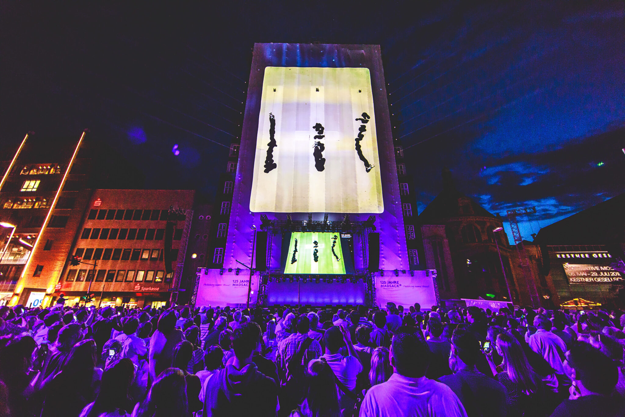 Night view across a packed city square, with a tall building lit in pink showing a flame-like projection and beams of light fanning out above the stage.
