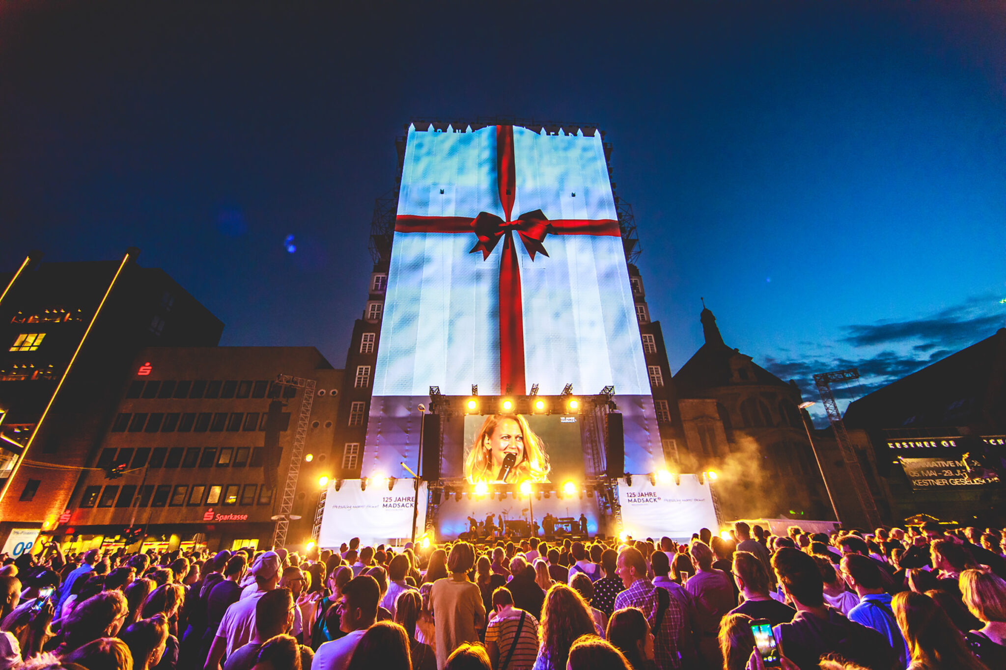 A tall building at dusk covered by a projection of white wrapping paper tied with a large red ribbon and bow, above a lit stage and a dense crowd.
