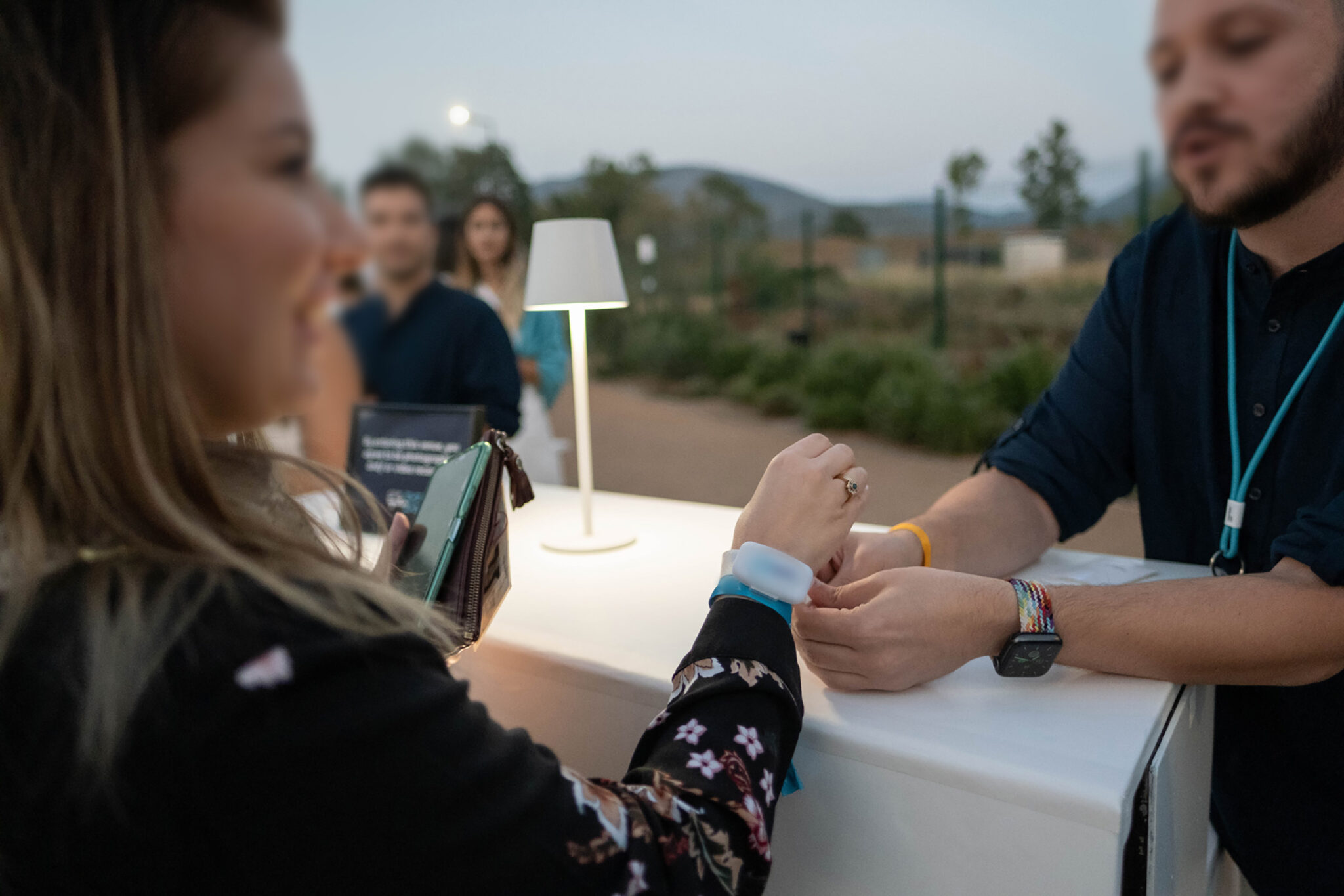A guest at a white counter holds out her phone to a staff member at dusk, with a small table lamp beside them.