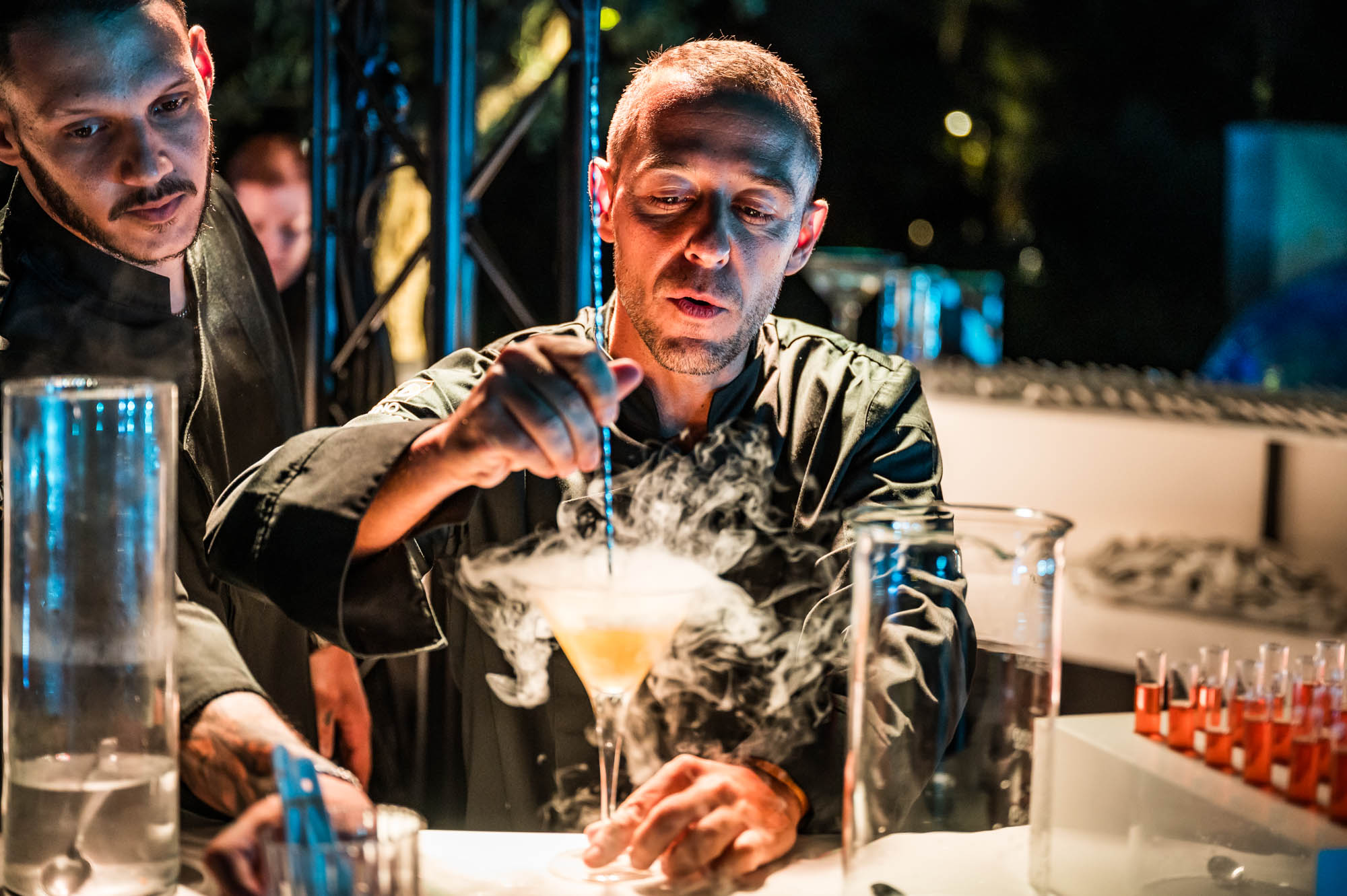 A bartender in black finishes a cocktail as smoke rises from the glass, with a second bartender watching behind him.