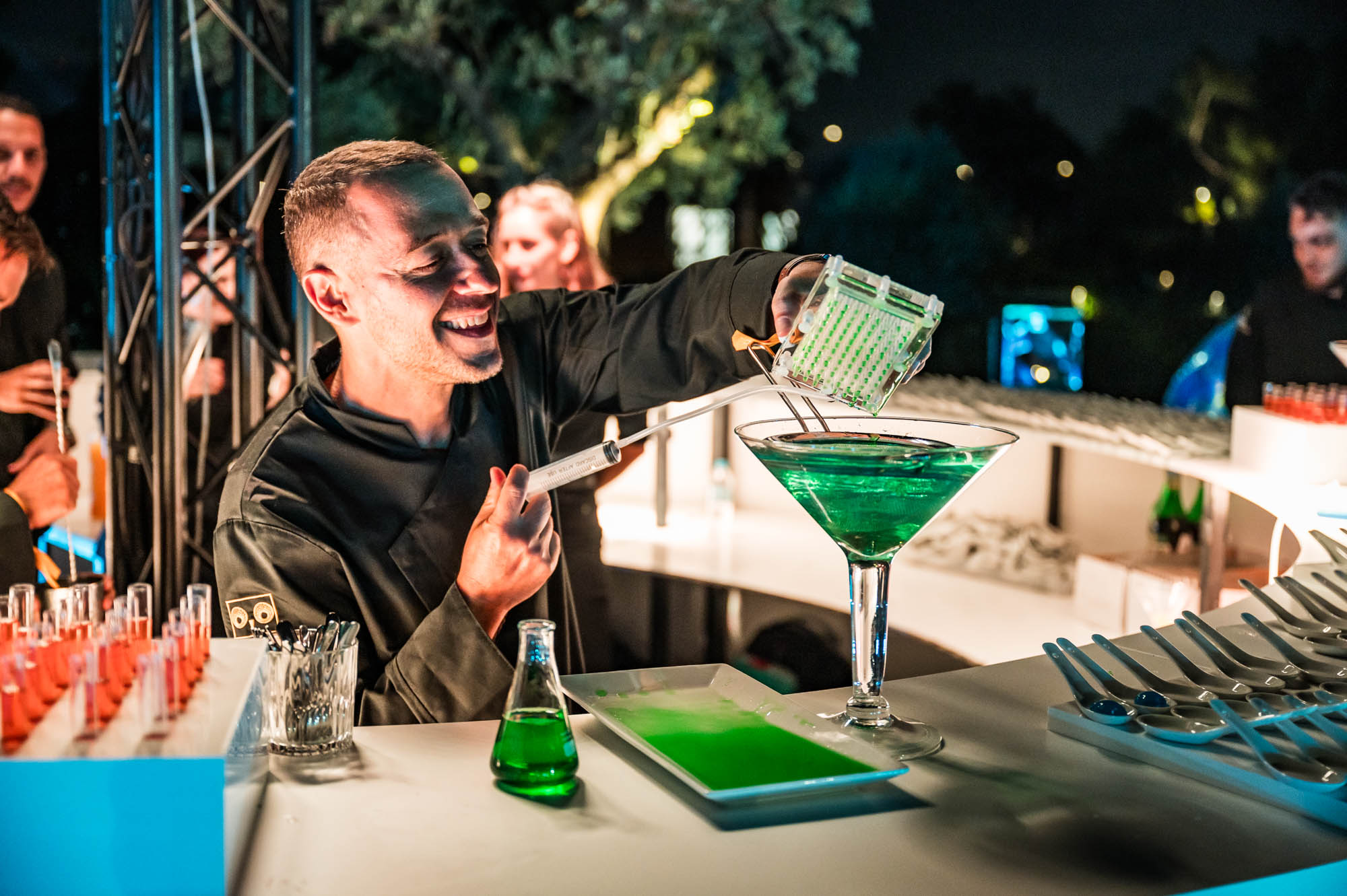 A bartender in black smiles while pouring a bright green drink into a cocktail glass, surrounded by glassware on a lit bar.