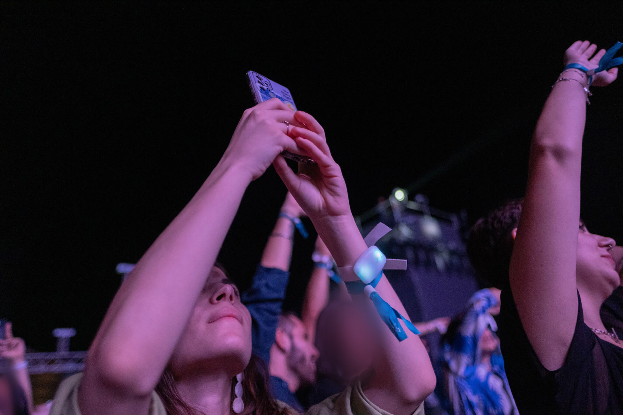Women in a crowd at night raise their arms to take photos with their phones, wearing event wristbands.
