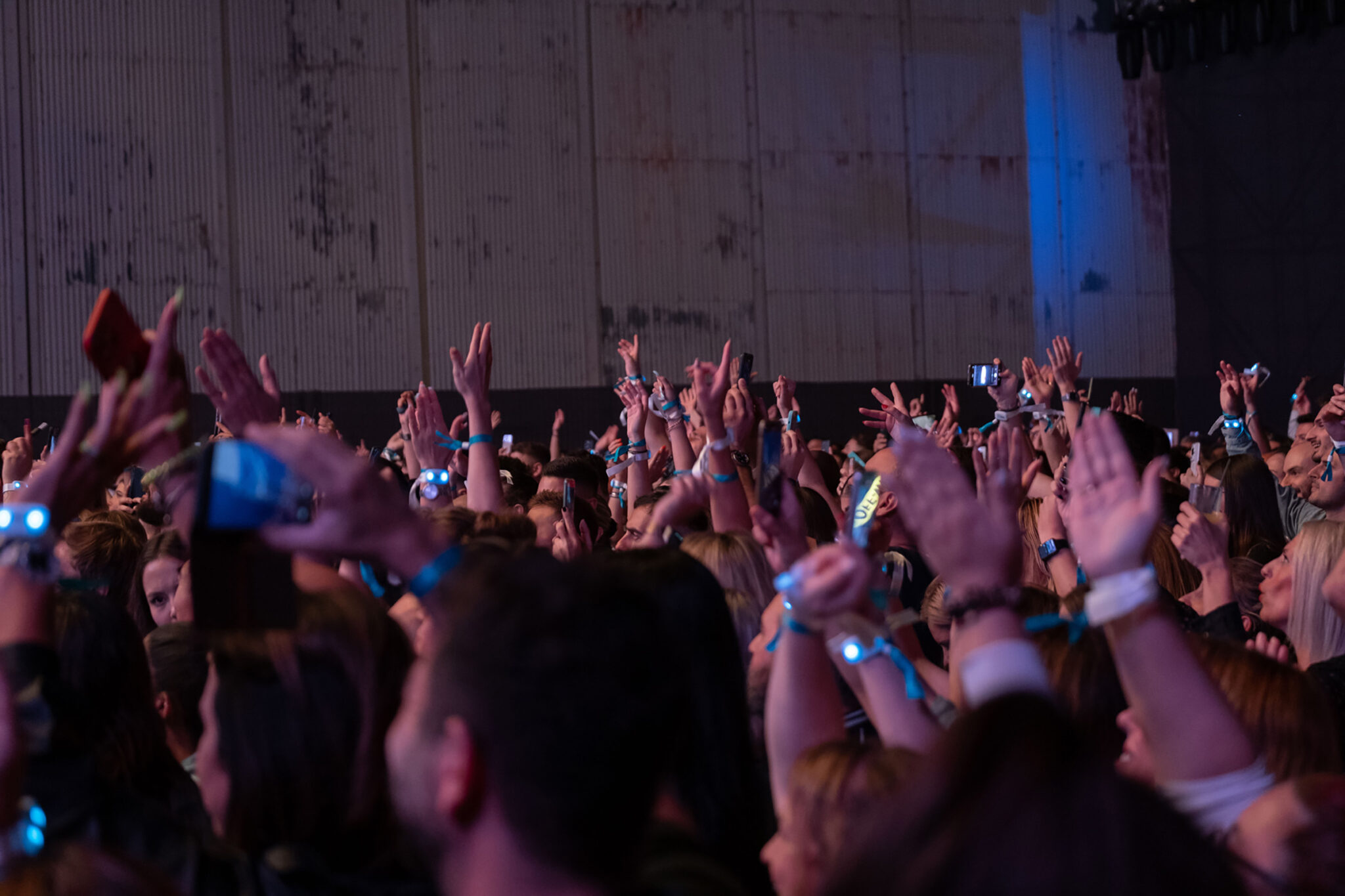 A dense crowd raises their hands and phones in front of a pale wall during a night performance.