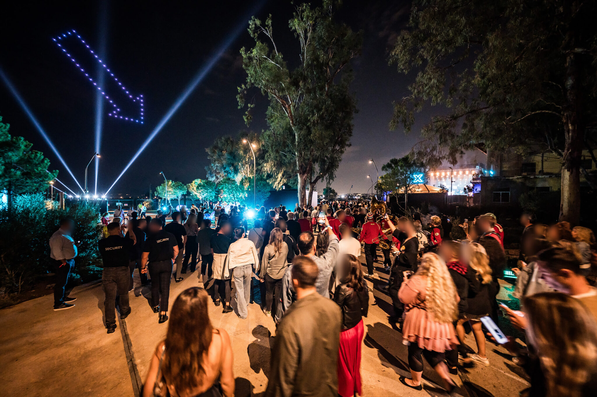 Guests walk along a tree-lined path at night as drones form a glowing arrow in the sky above them.