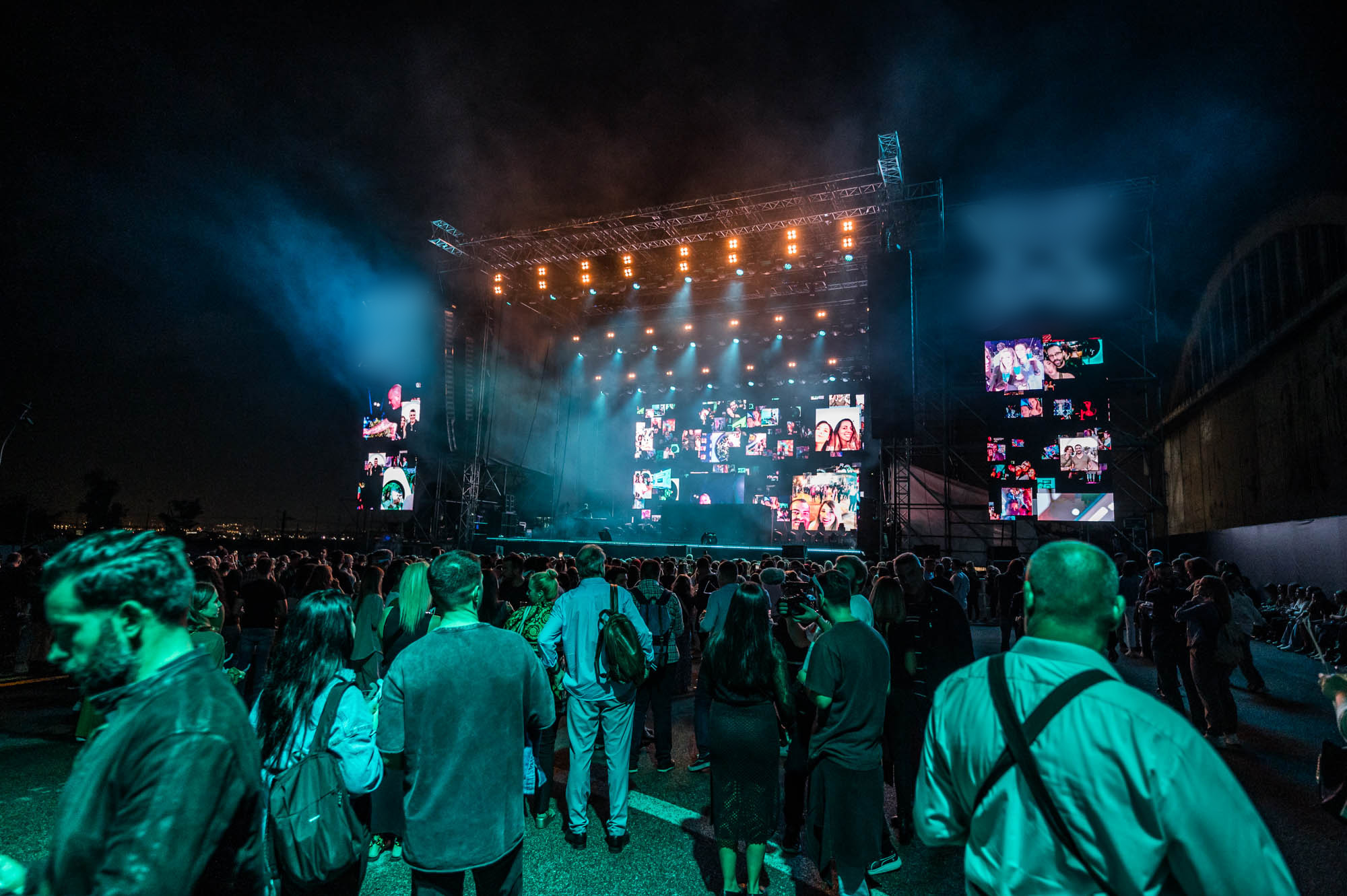 A crowd stands in front of an outdoor stage with large screens and teal haze, some guests in the foreground lit in green.