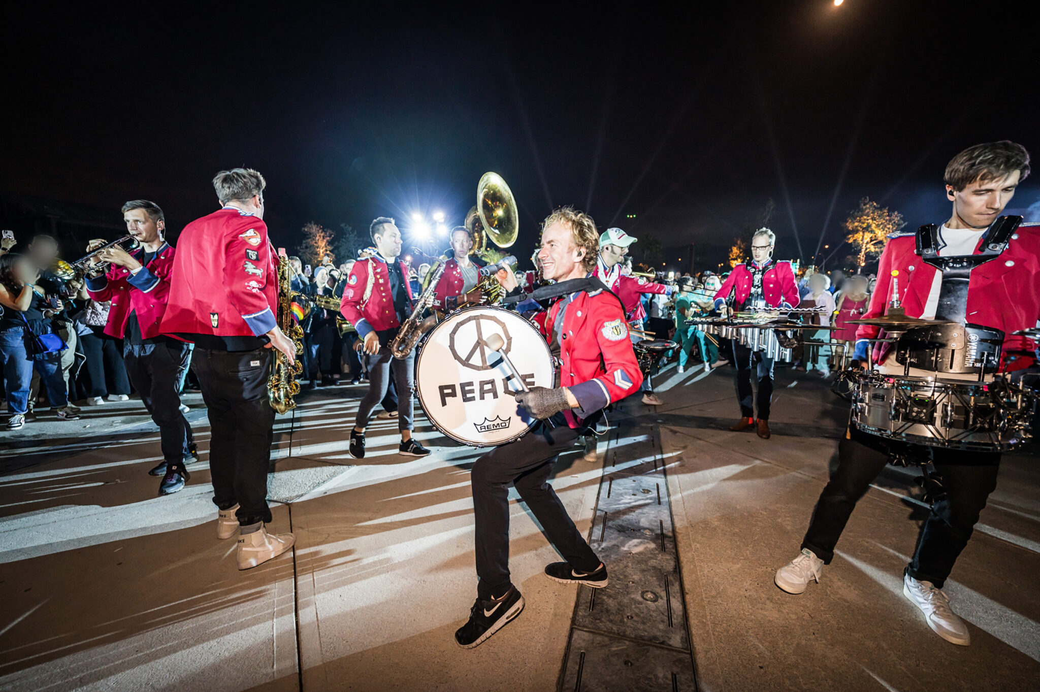 A marching band in red jackets plays drums and brass instruments while walking through the crowd at night.