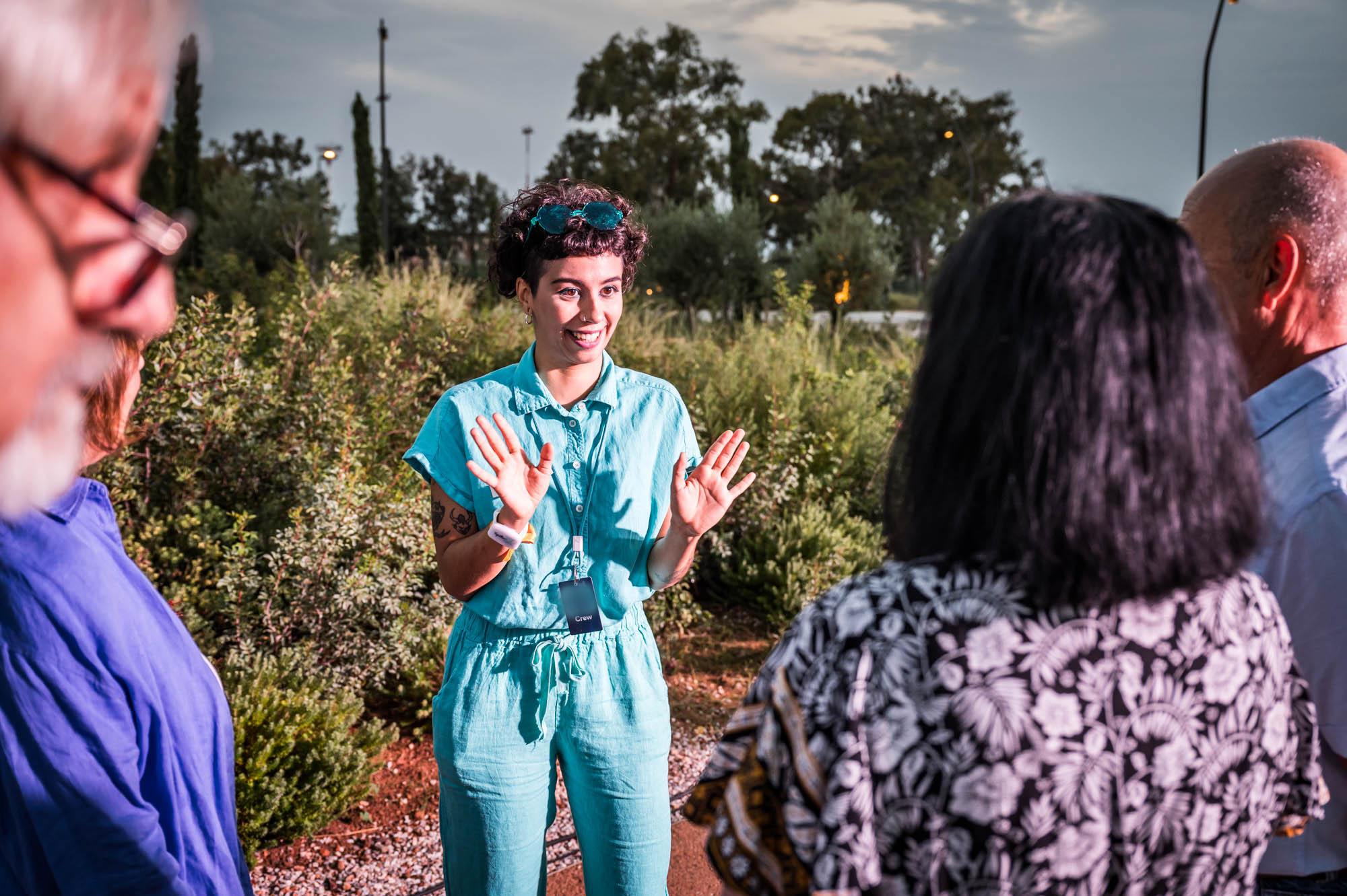 A host in a turquoise jumpsuit smiles and gestures while welcoming a small group of guests outdoors at dusk among green shrubs.
