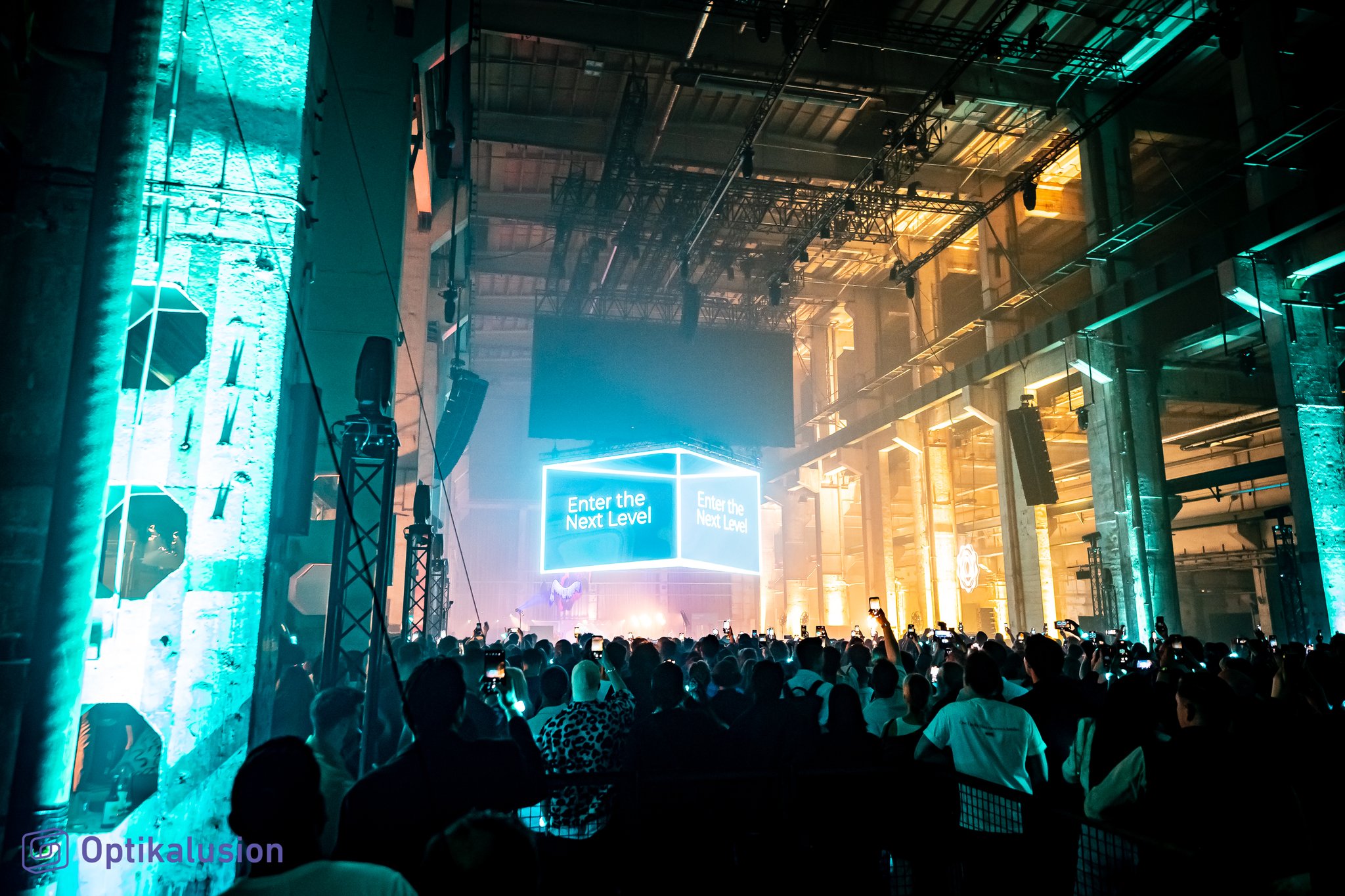 A large audience faces a stage with a glowing screen at the far end of a long industrial hall, framed by a tall teal light column.