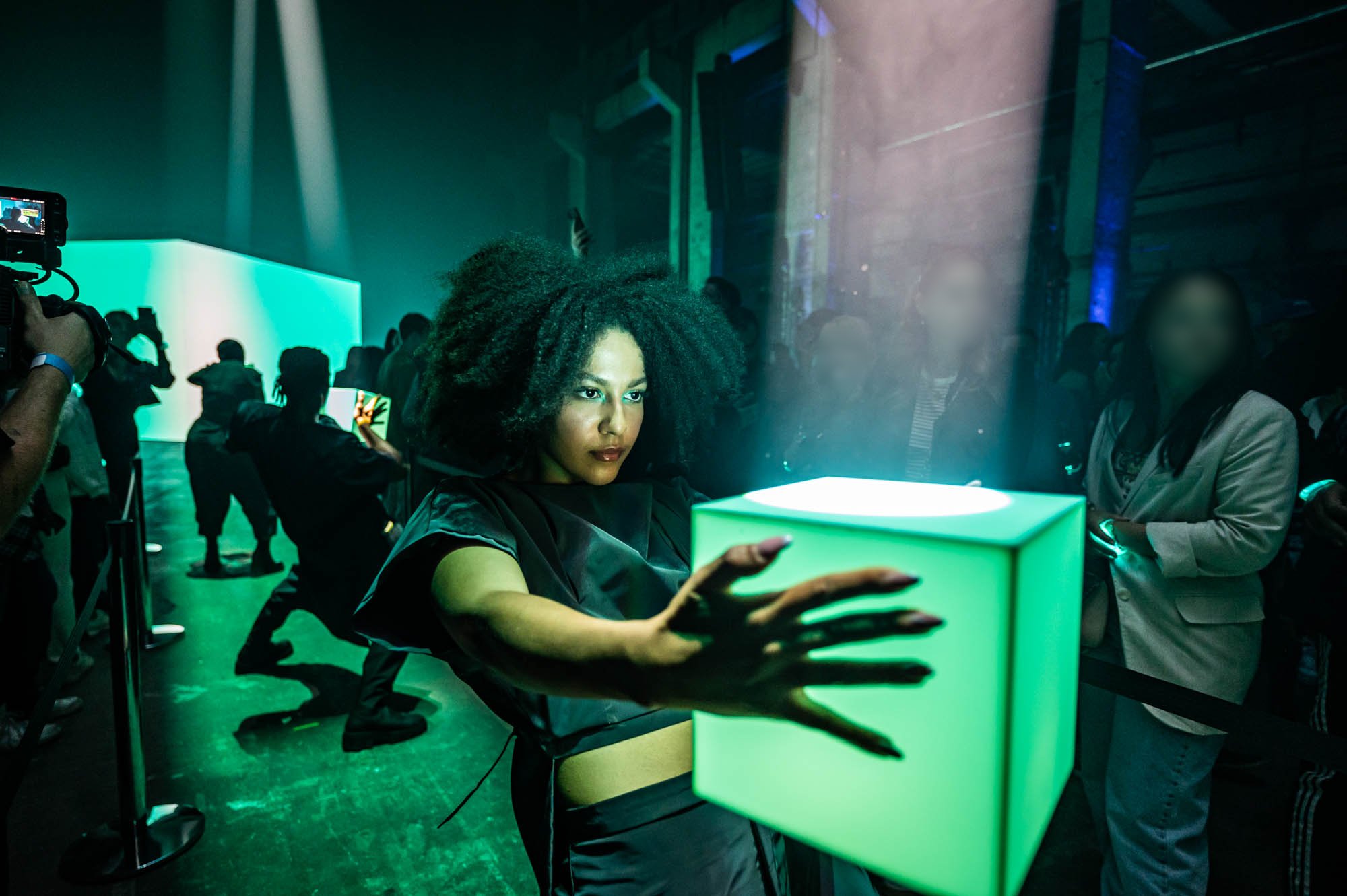 A performer reaches toward a glowing green cube on a pedestal, with other performers and guests in a teal-lit hall behind her.
