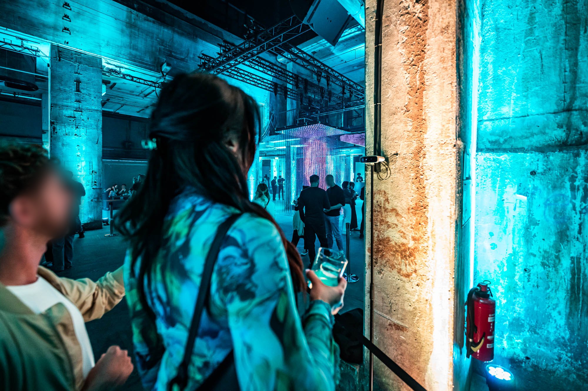 A guest with long hair walks past a concrete pillar into a hall lit in teal, with other guests nearby.