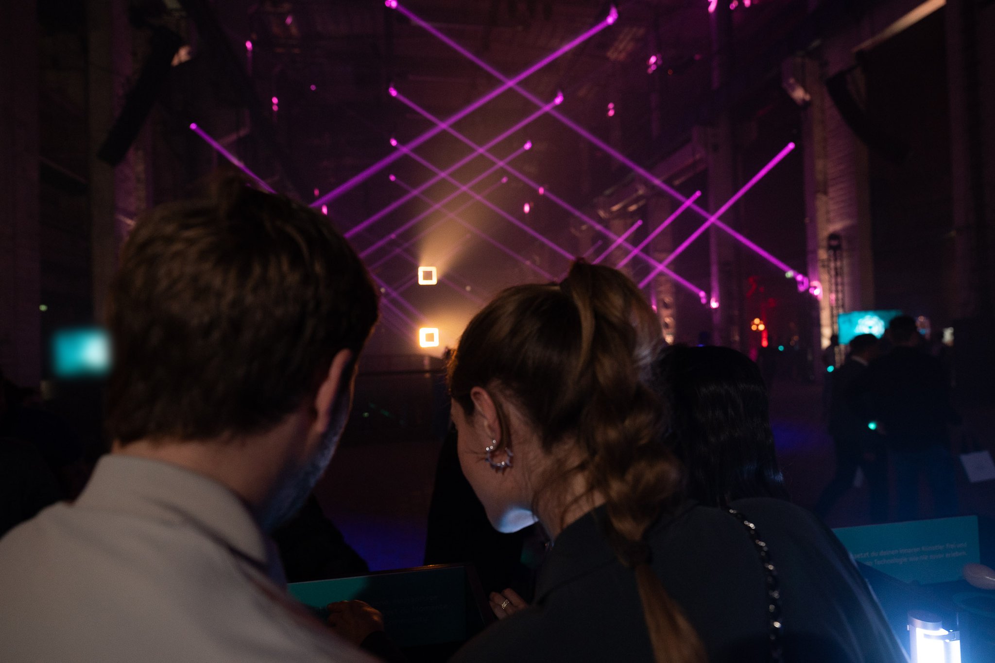 Two guests seen from behind watch purple and white laser beams sweep across a dark hall.