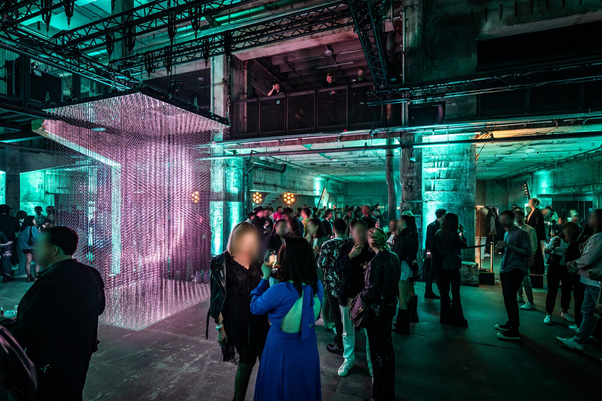 Guests stand and talk in a large industrial hall beside a tall installation of hanging pink light strands.