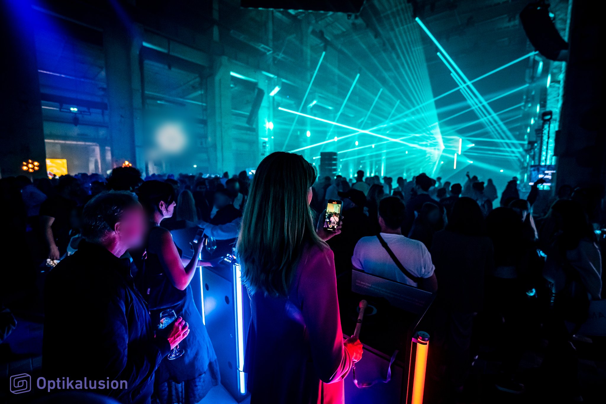 Guests in the foreground watch teal laser beams and haze fill a hall packed with people.