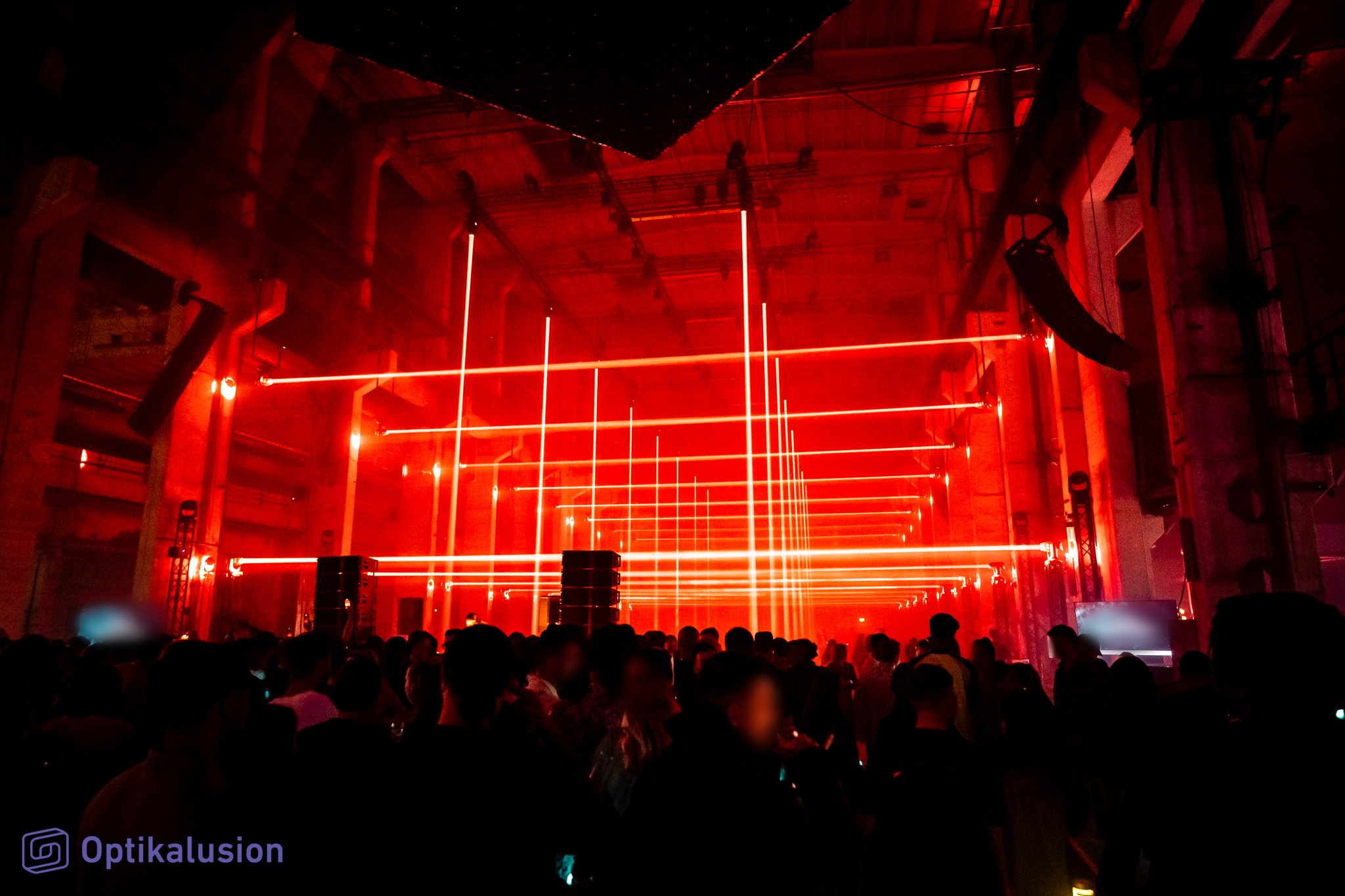 A grid of red light lines forms a glowing tunnel above a crowd in a dark industrial hall.