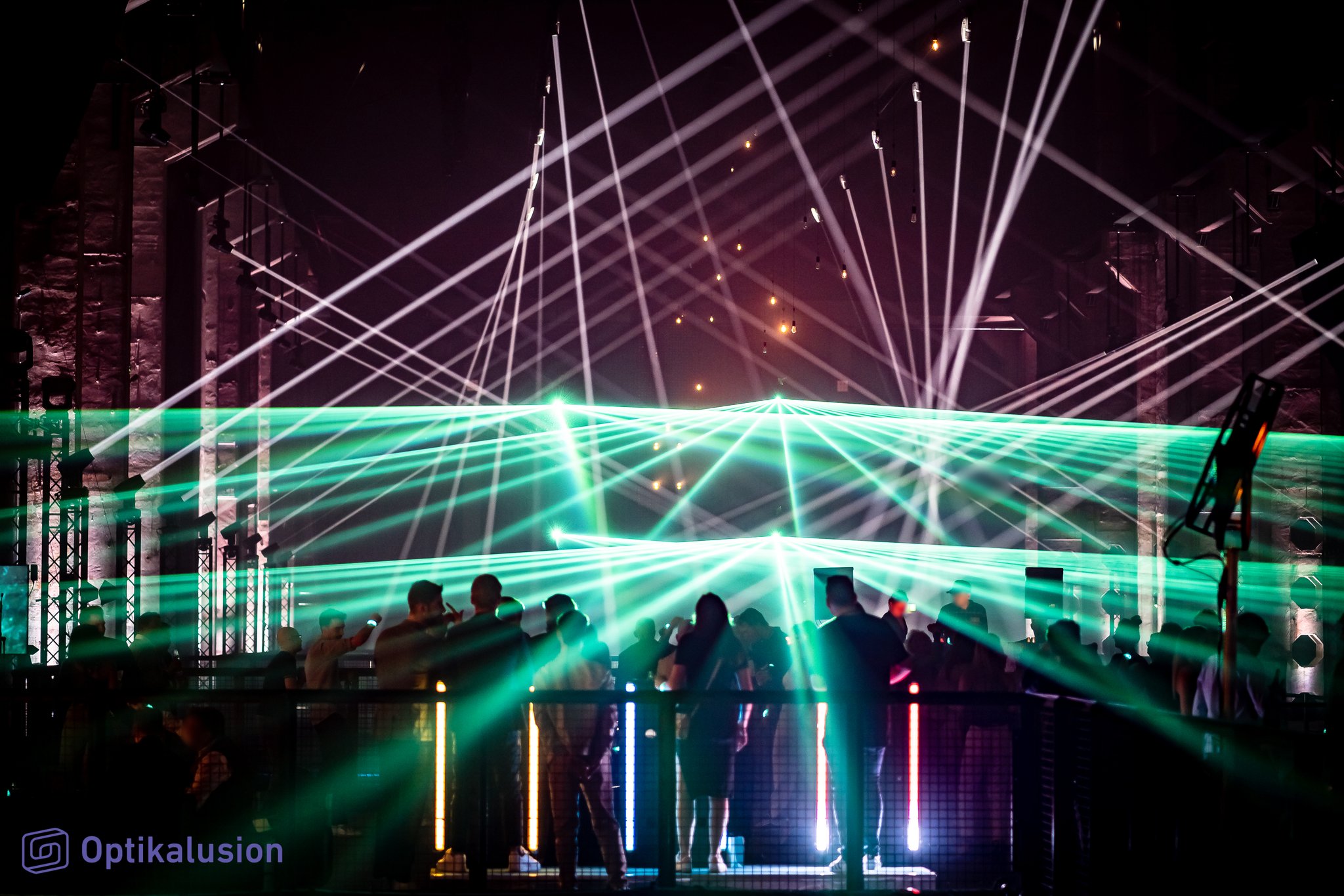 White and green laser beams sweep across a hall above a crowd and a lit bar on a raised level.