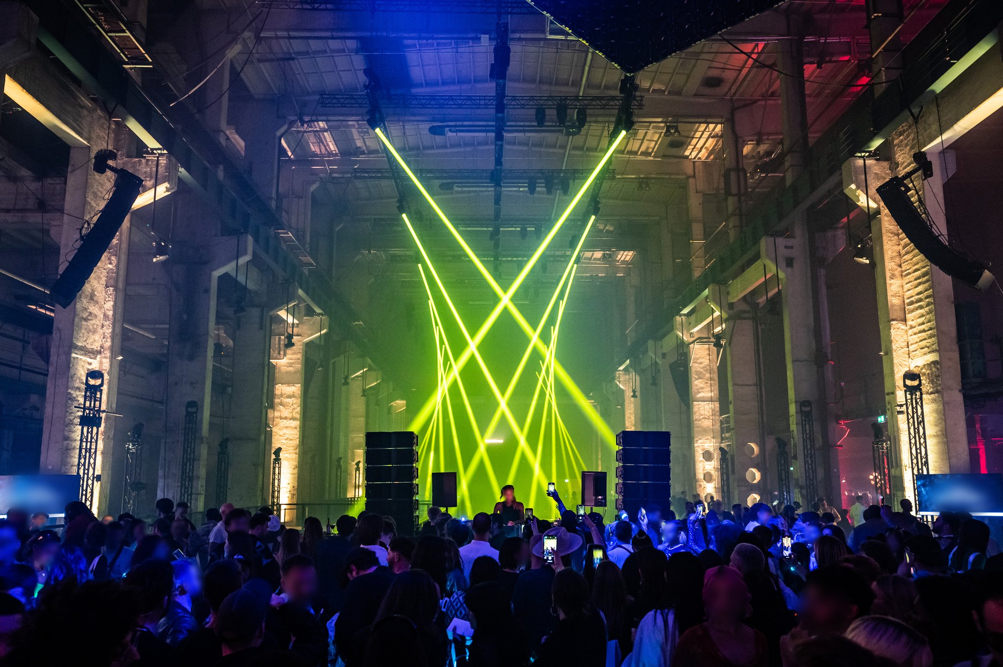 A crowd stands beneath crossing yellow-green laser beams in a tall industrial hall with stacked speakers on stage.